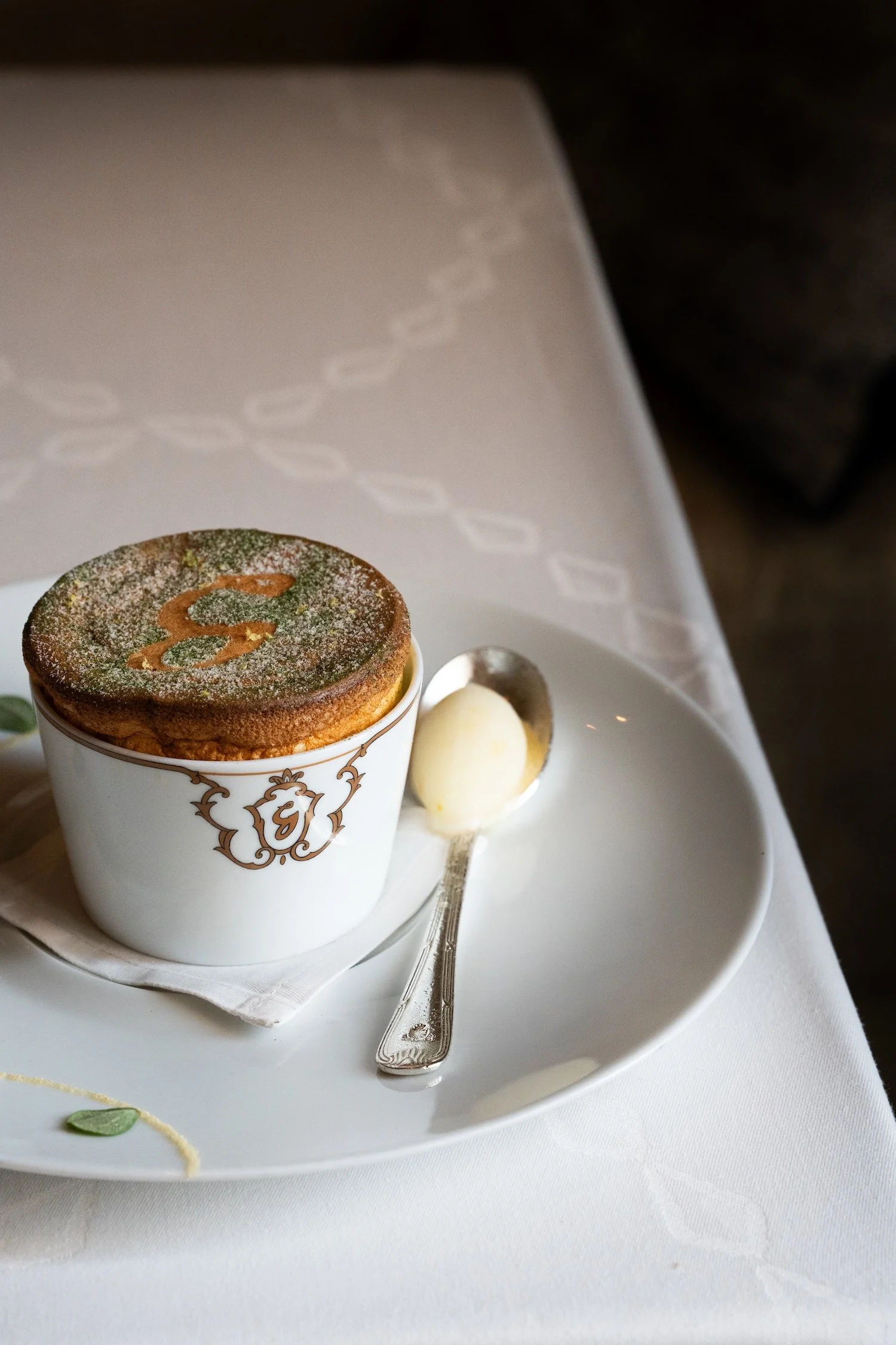 Un dessert dans un petit pot blanc avec un logo ornemental, saupoudré de poudre de cacao et décoré d'un motif en forme de sur le dessus. Accompagné d'une boule de glace ou de sorbet sur une assiette blanche, une cuillère en argent. Le tout présenté sur un plateau blanc avec une nappe blanche.