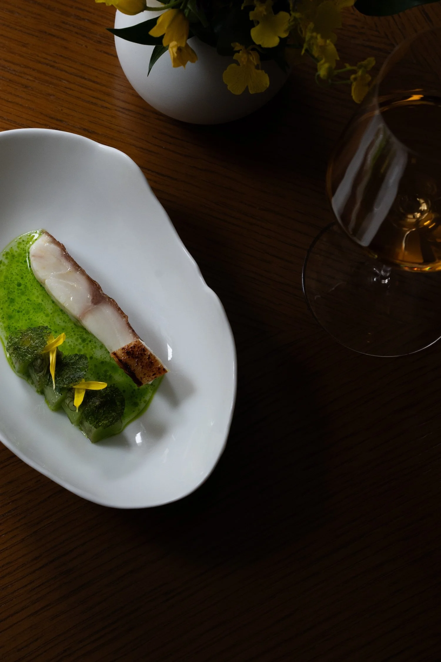 Une assiette blanche avec un plat de poisson, accompagné d'une sauce verte, décoré de fleurs jaunes et de feuilles. Un verre de vin blanc et un vase avec des fleurs jaunes sont également visibles sur la table en bois.