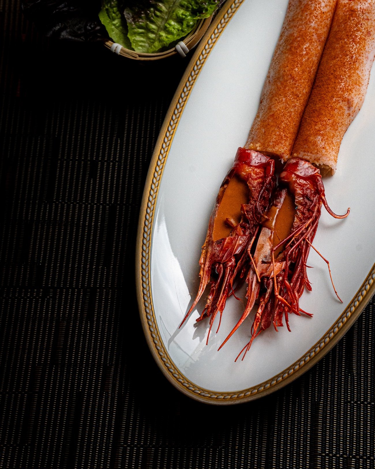 Crevettes rouges sur une assiette blanche dorée avec une sauce, accompagnées de laitue en arrière-plan.