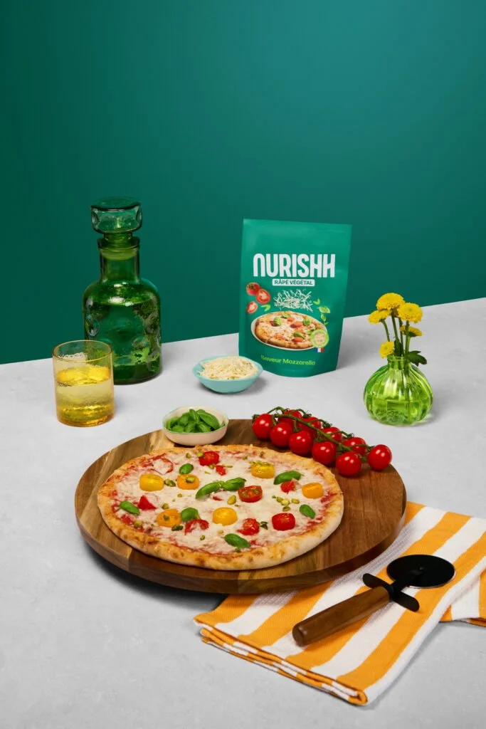 Une pizza garnie de tomates cerises, basilic, et autres légumes, sur une planche en bois. À côté, des tomates cerises en grappes, un bol de basilic frais, un verre de boisson jaune, et un petit bol de fromage râpé. En arrière-plan, un paquet de Nourish végétal, une bouteille verte et un verre. Sur la droite, un vase avec des fleurs jaunes et un torchon blanc à rayures jaunes, avec un ouvre-boîte noir et en bois.