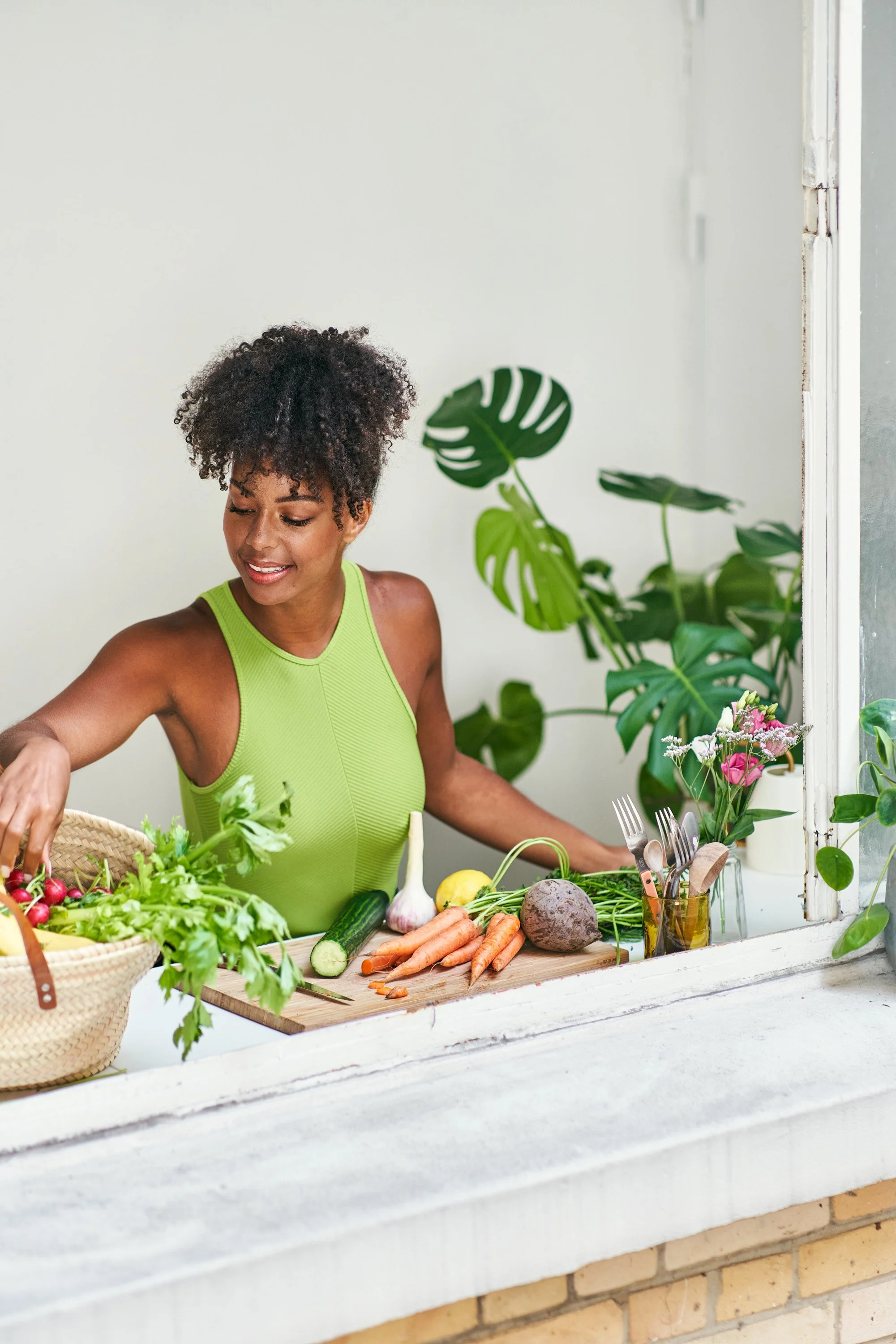 Une femme souriante en haut d'un balcon, préparant des légumes frais pour un repas, entourée de plantes vertes et de fleurs.