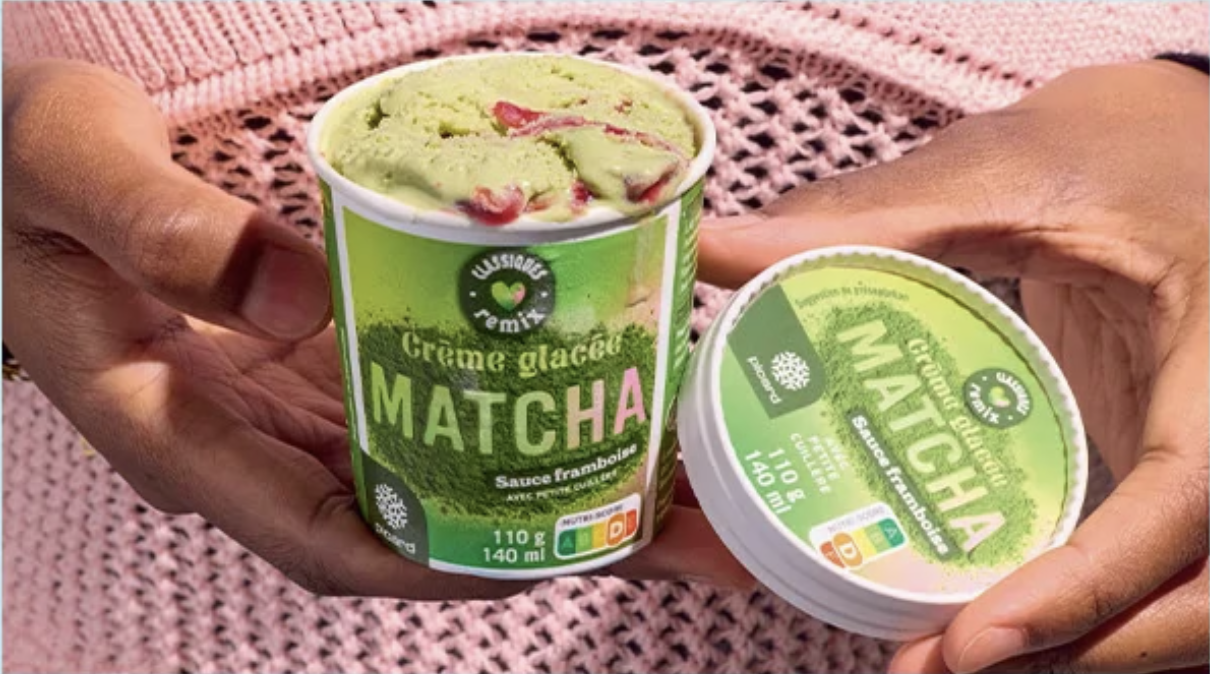 Une personne tient un pot de crème glacée matcha avec une sauce aux framboises visible à l'intérieur, sur un fond de surface en crochet rose.