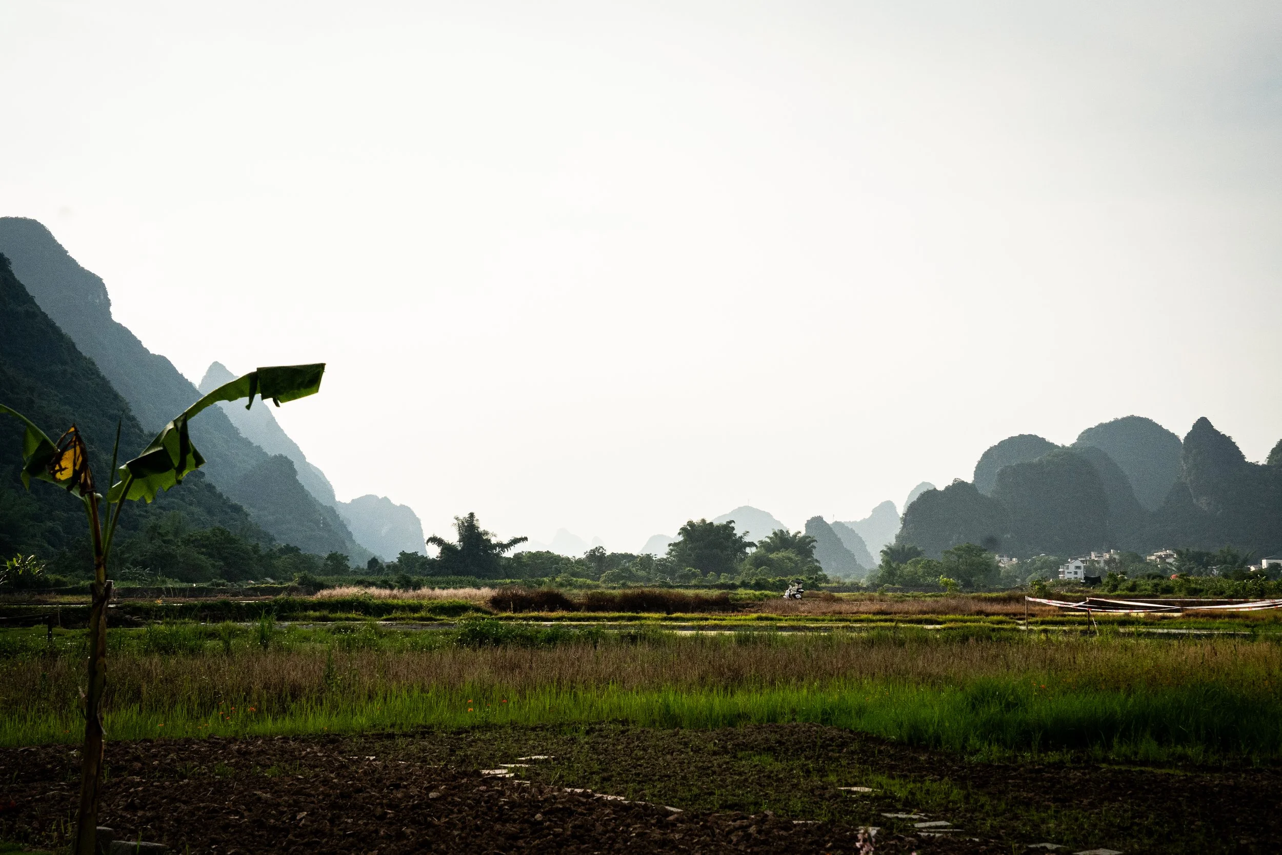Paysage rural avec des montagnes verdoyantes, un champ cultivé, quelques bâtiments et un scooter à l'horizon, par une journée ensoleillée ou légèrement nuageuse