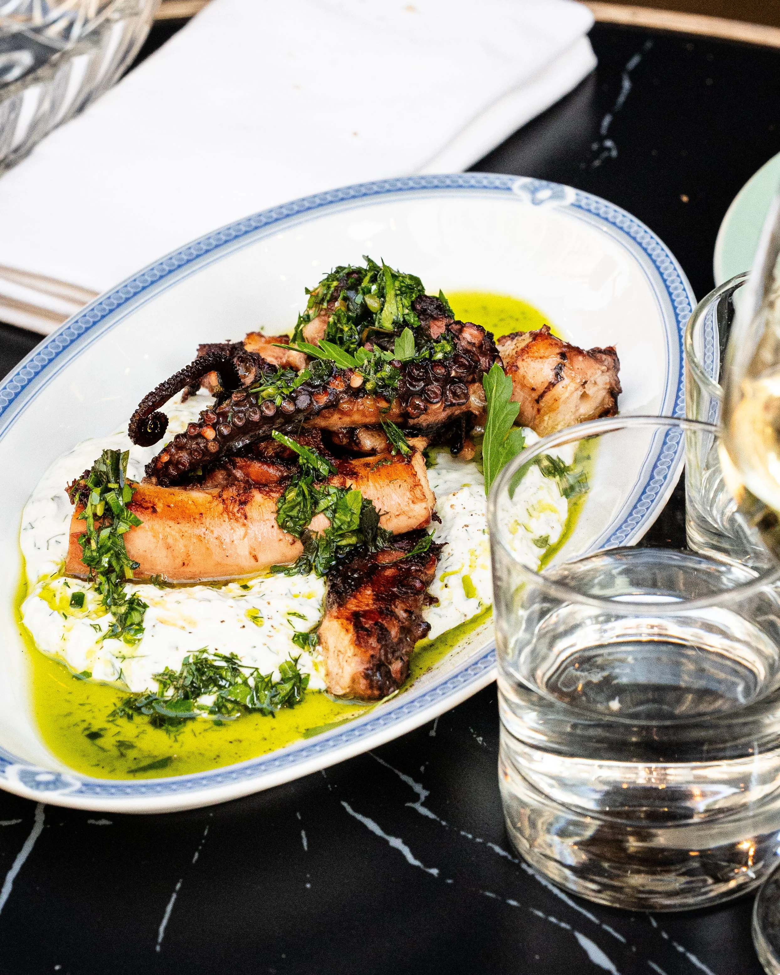 Plat de poisson grillé avec légumes verts, sauce verte, et huile d'olive, servi dans un plat en céramique blanche avec bord bleu, à côté de verres d'eau.
