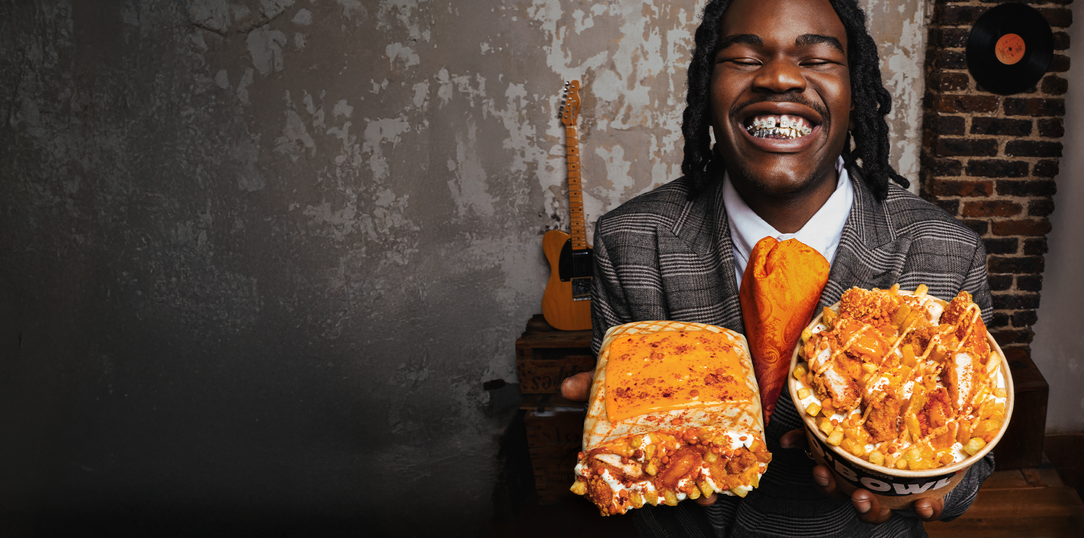 Homme souriant tenant deux plats de nourriture, un gâteau et un bol de poulet frit avec frites et sauce, en intérieur avec guitare et mur en brique en arrière-plan.