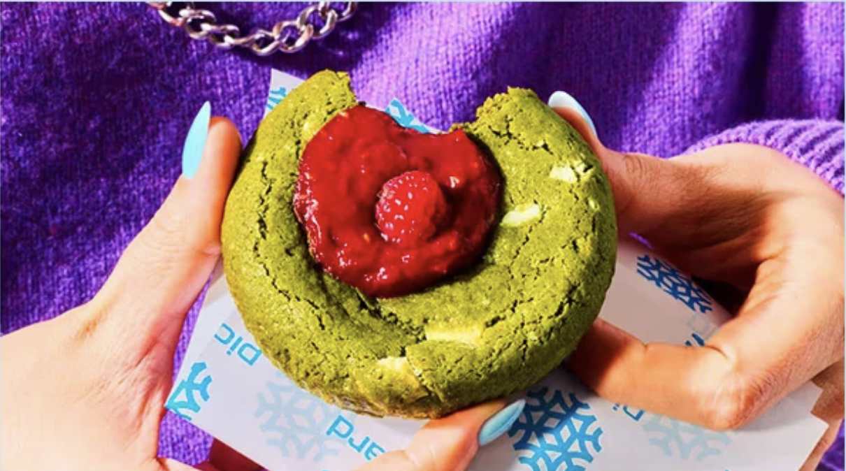 Une personne tient un muffin vert avec une garniture rouge à la fraise et une fraise en décoration, sur une nappe blanche avec des motifs de flocons de neige bleus.