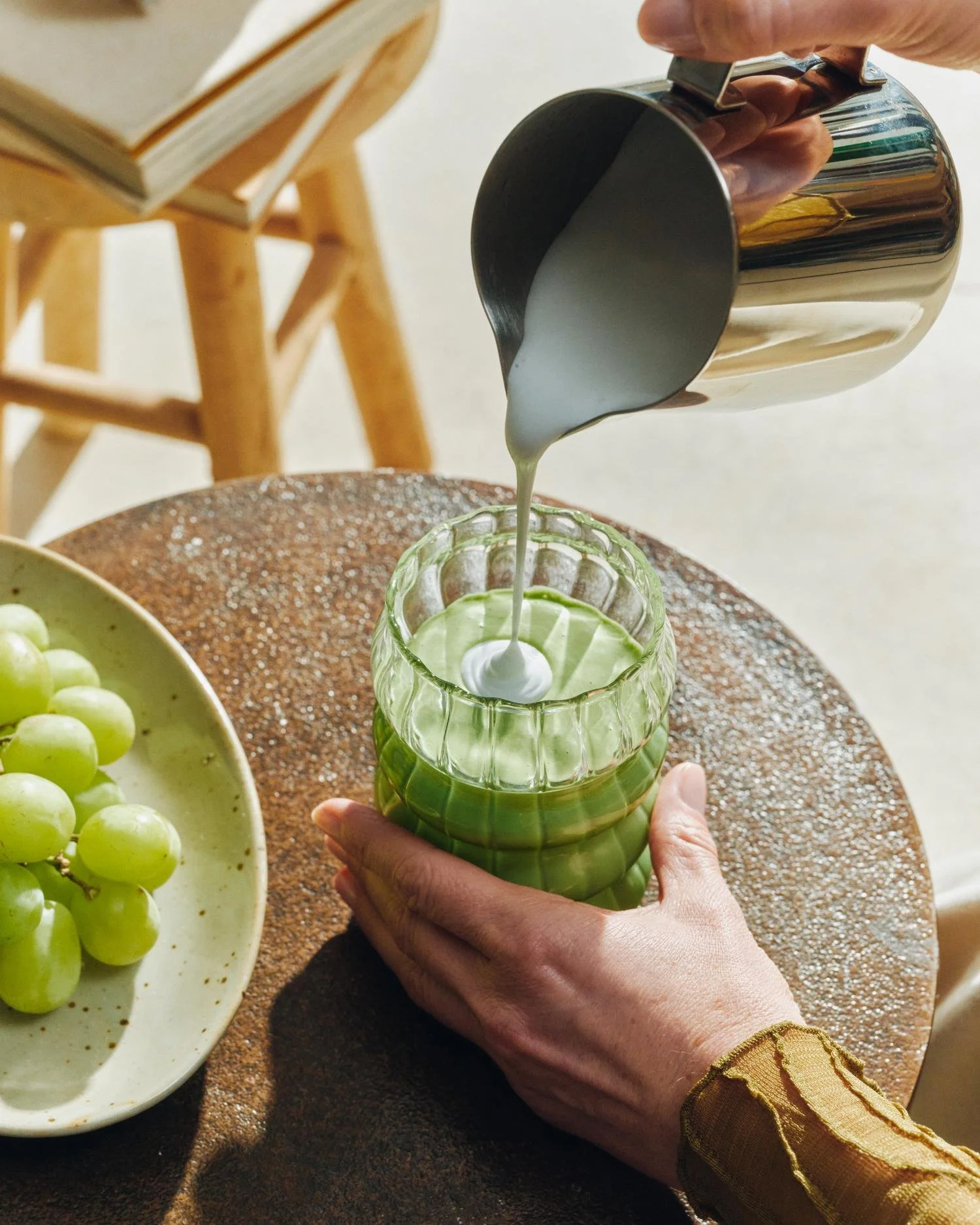Une personne verse du lait dans un verre en verre vert avec une décoration en forme de fleurs, avec une panière de raisin vert à côté, sur une table en bois.