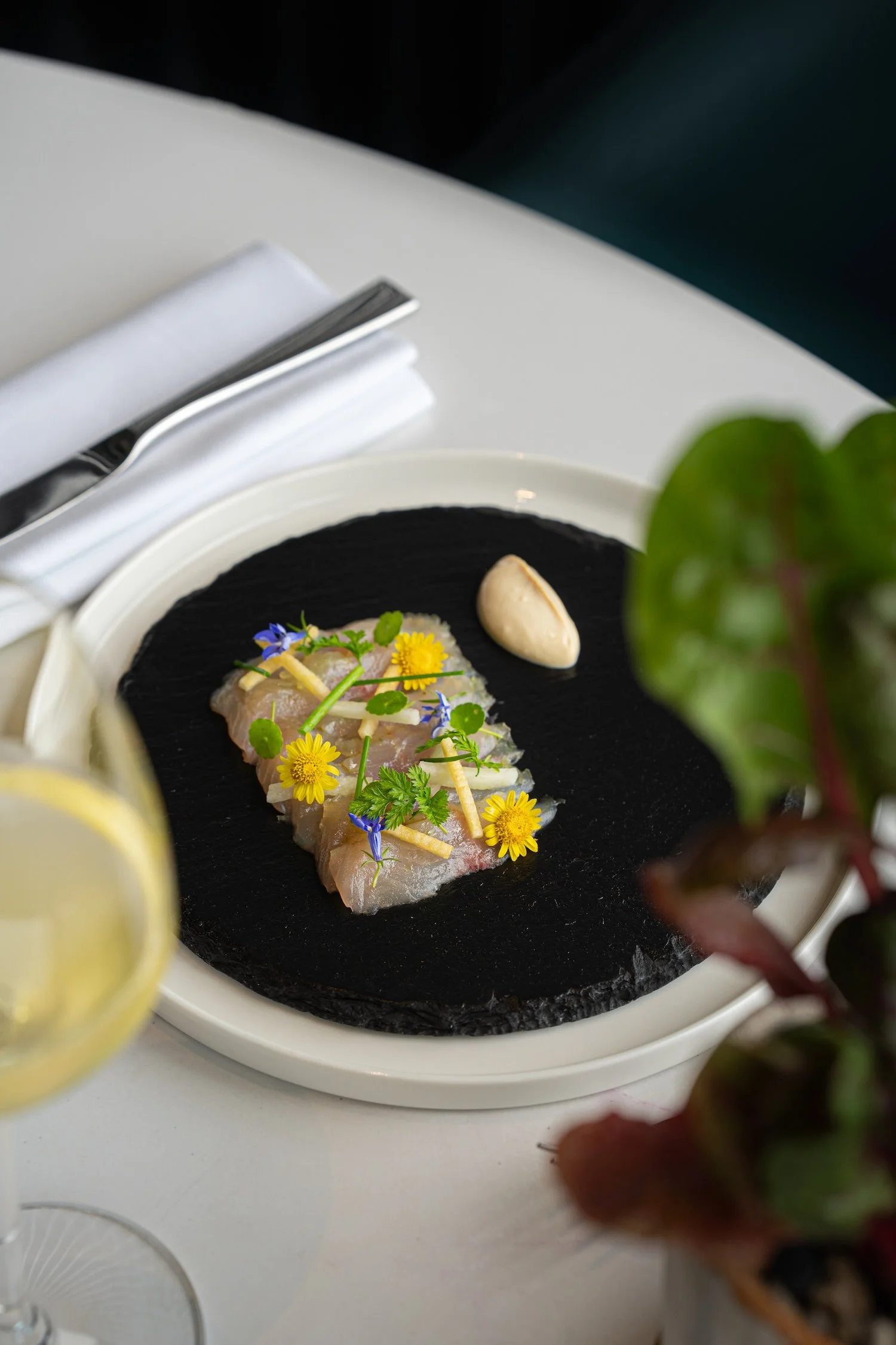 Assiette de poisson cru garni de fleurs comestibles, accompagnée d'une tranche de citron et d'un condiment, sur un fond blanc avec un couvert à droite