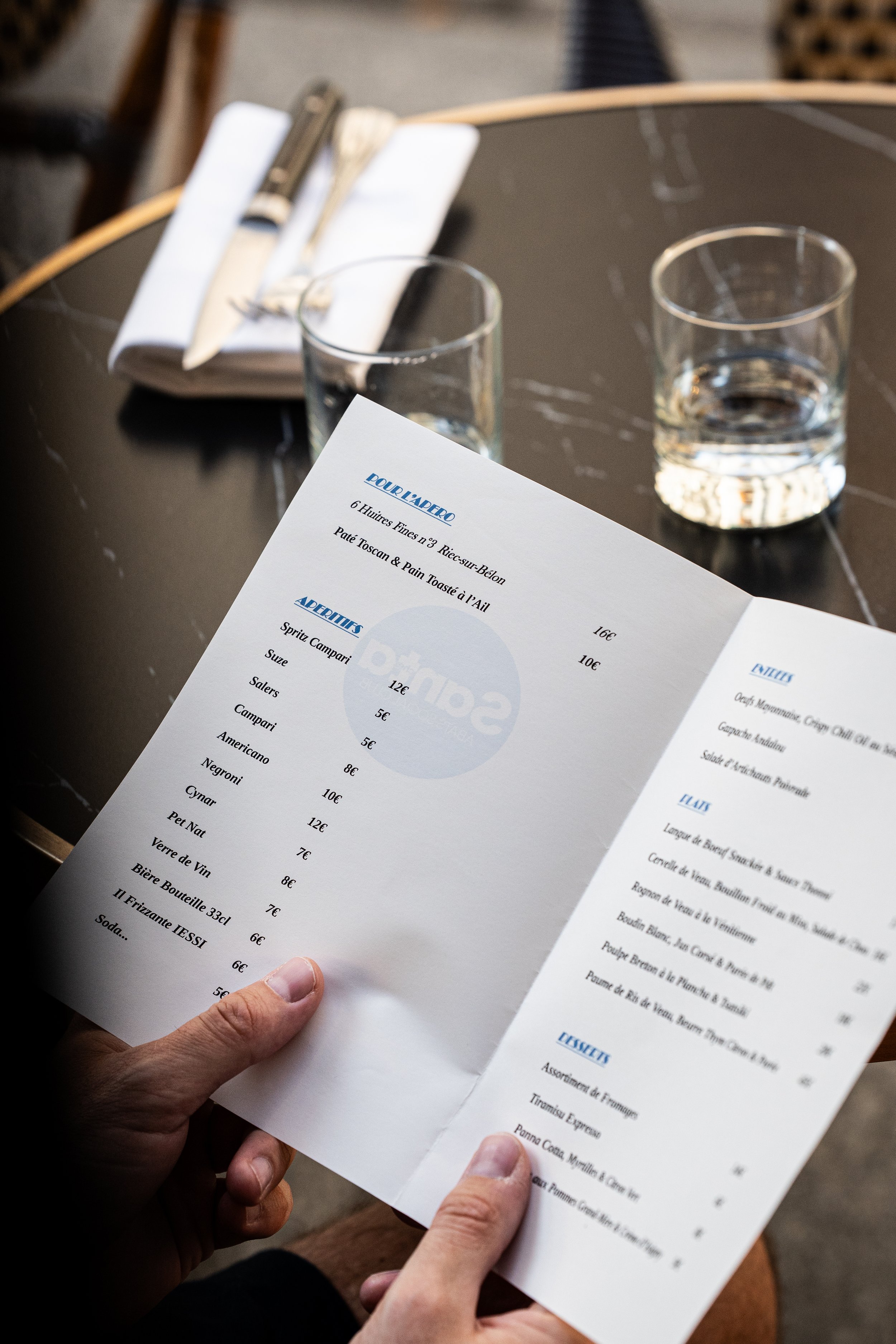 Une personne tient une menu de restaurant en main, avec des verres d'eau sur la table et des couverts posés sur une serviette.