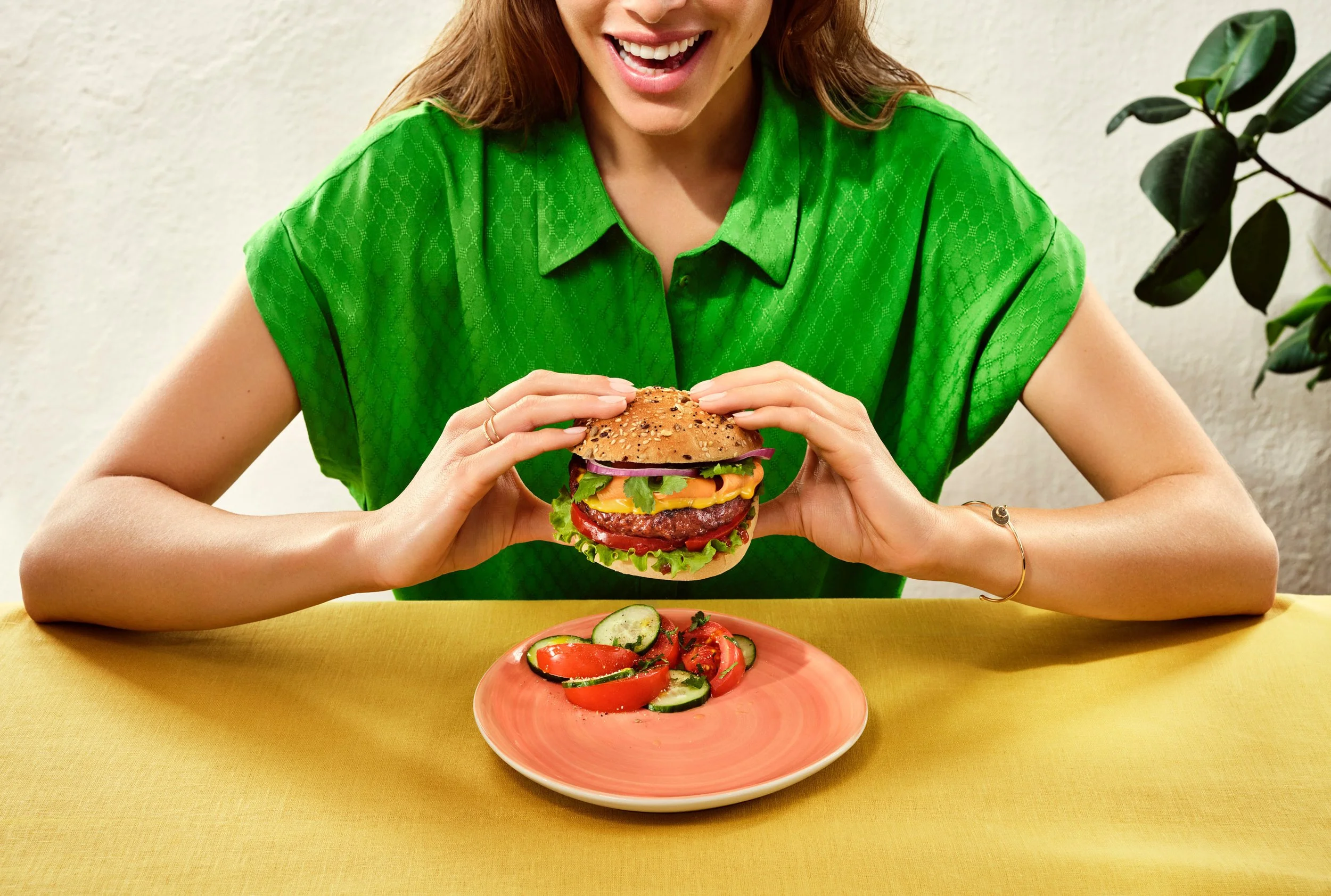 Une femme souriante en chemise verte tient un hamburger avec un pain aux graines. Il y a une assiette de tomates et concombres tranchés devant elle et un plant de plante verte à l'arrière.