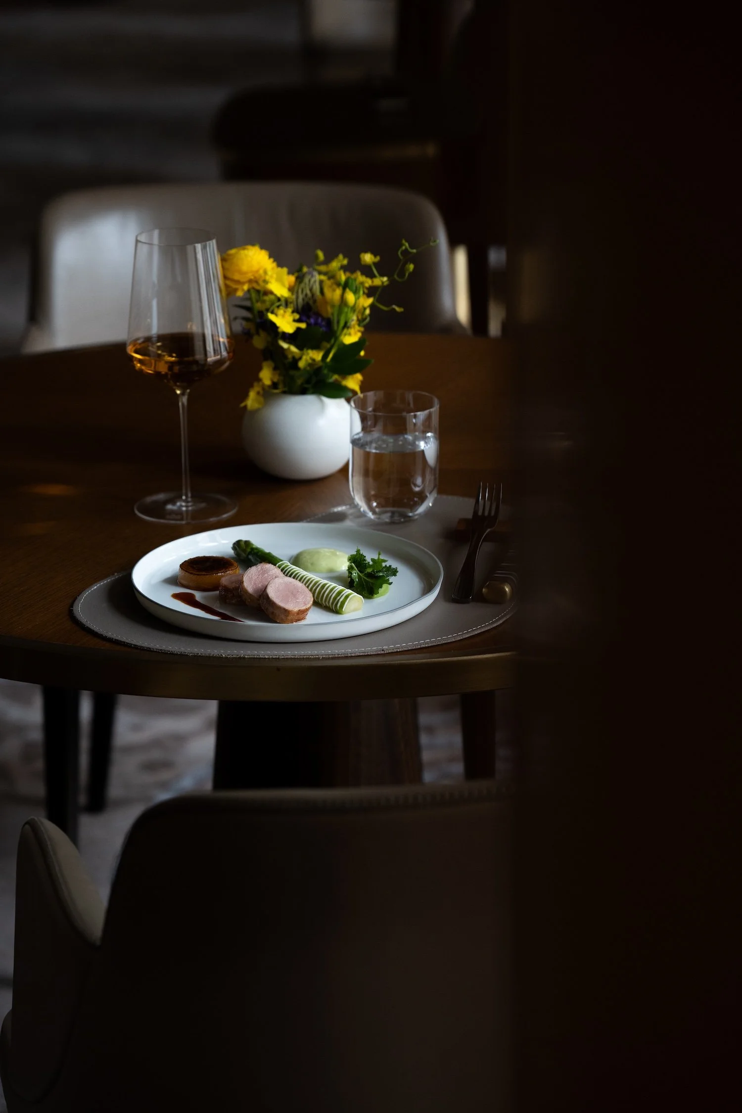 Table avec un verre de vin, un verre d'eau, un plat avec de la viande, une sauce et des légumes, une petite bouteille de sauce, une cuillère, une fourchette, une vase avec des fleurs