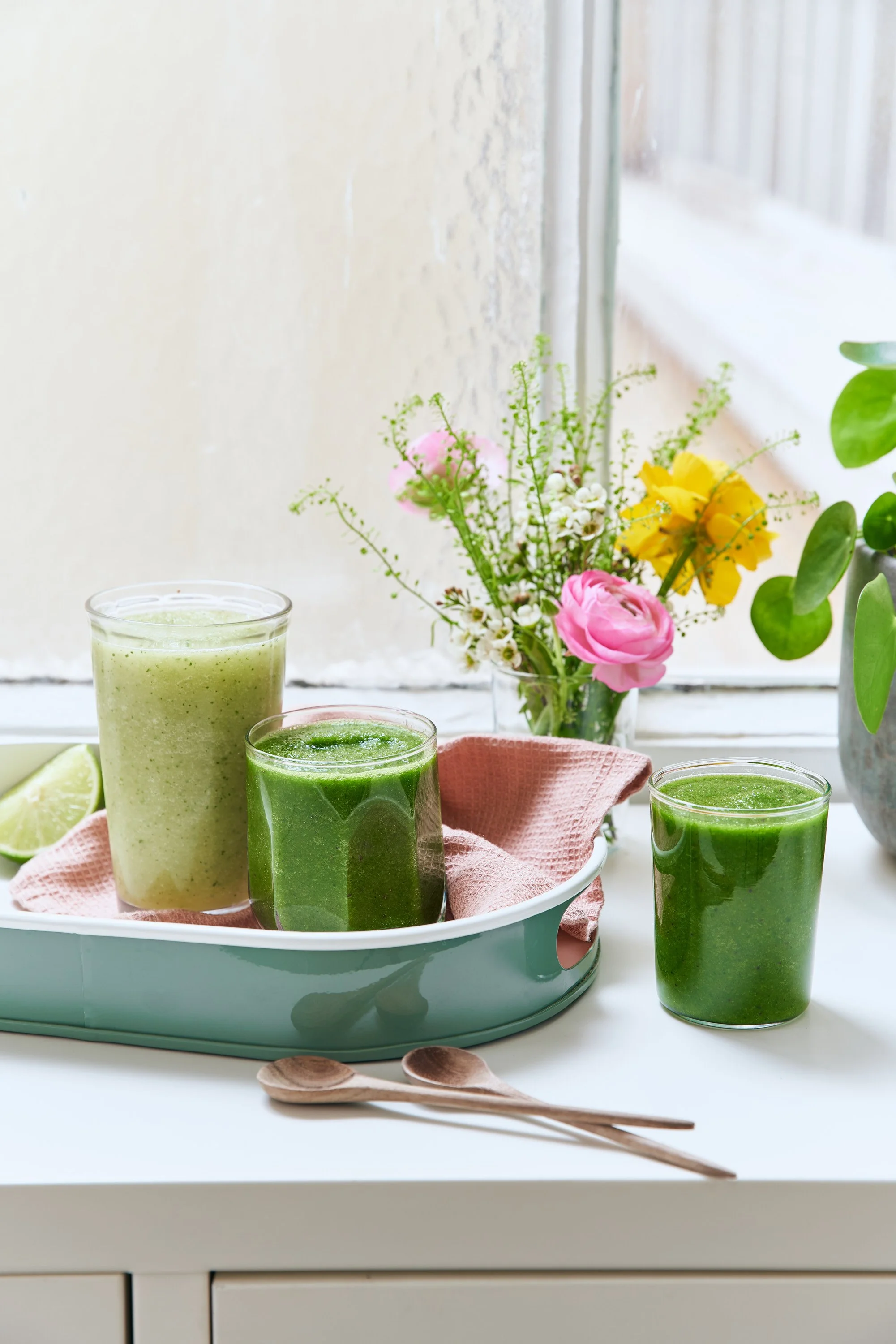 Verres de smoothies verts, une petite tartelette et deux cuillères en bois avec une écharpe en tissu rose clair, avec des fleurs dans un vase en arrière-plan sur une fenêtre
