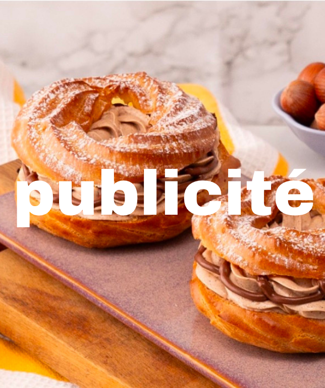 Deux petits choux garnis de crème au chocolat sur une planche en bois, avec un bol de noisettes en arrière-plan.
