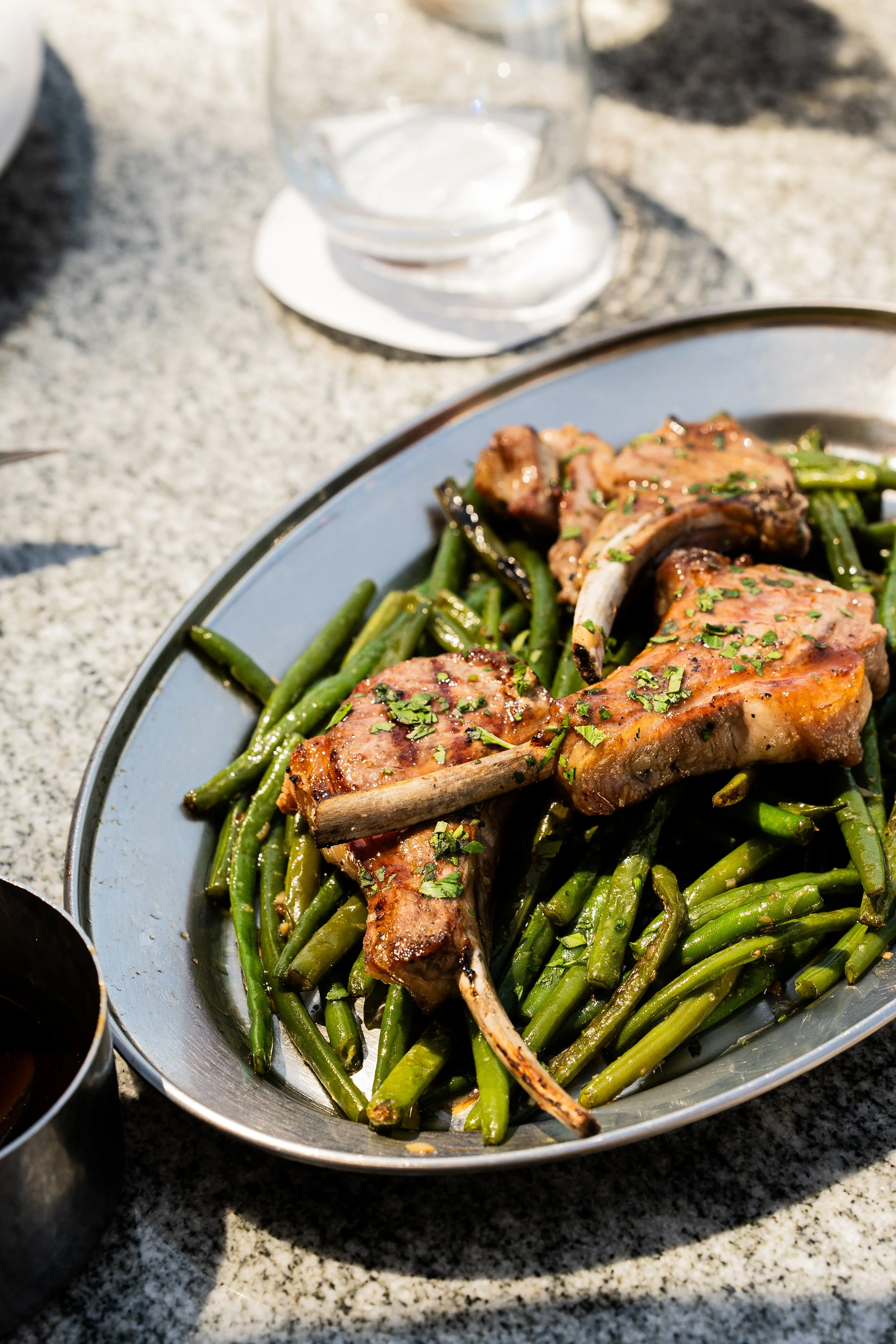 Une assiette avec des côtelettes d'agneau grillées et des haricots verts, sur une table en pierre, accompagnée d'un verre d'eau.