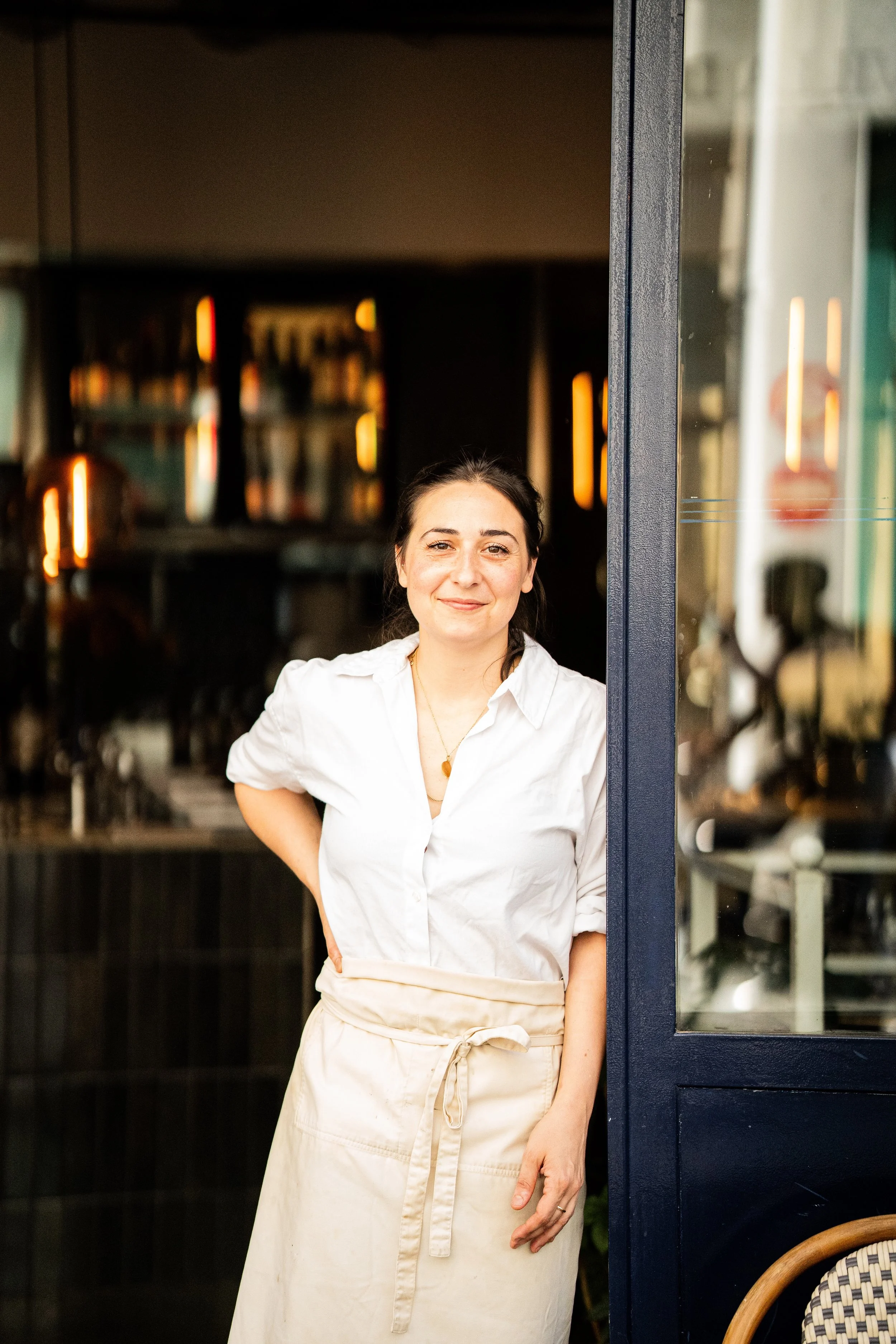 Une femme souriante portant une chemise blanche et un tablier beige, debout à la porte d’un restaurant ou d’un café, avec un fond flou et une ambiance chaleureuse.