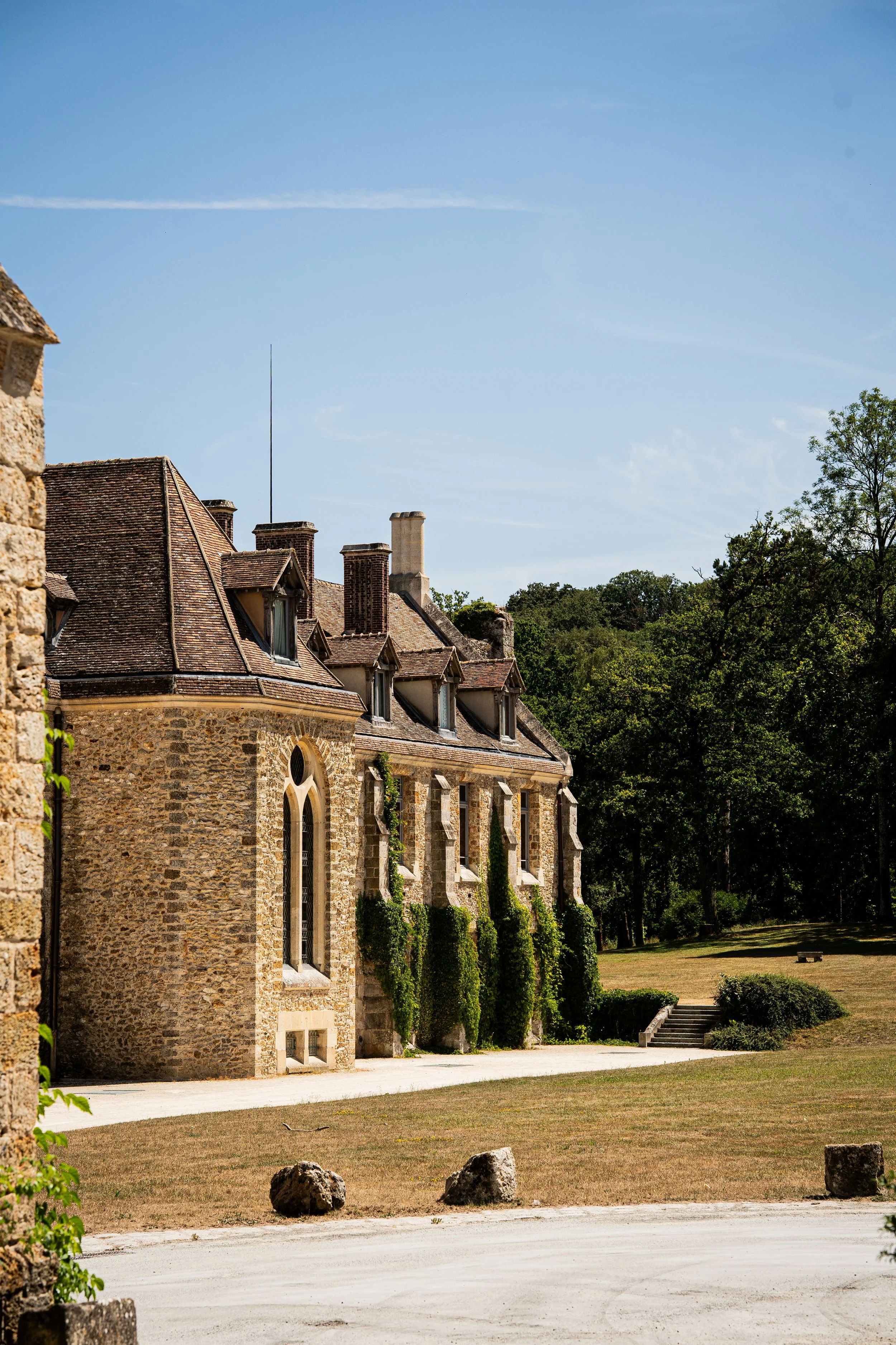 Vue d'un bâtiment en pierre avec un toit en tuiles, surplombant un espace de pelouse avec quelques rochers et arbres en arrière-plan, sous un ciel bleu clair.