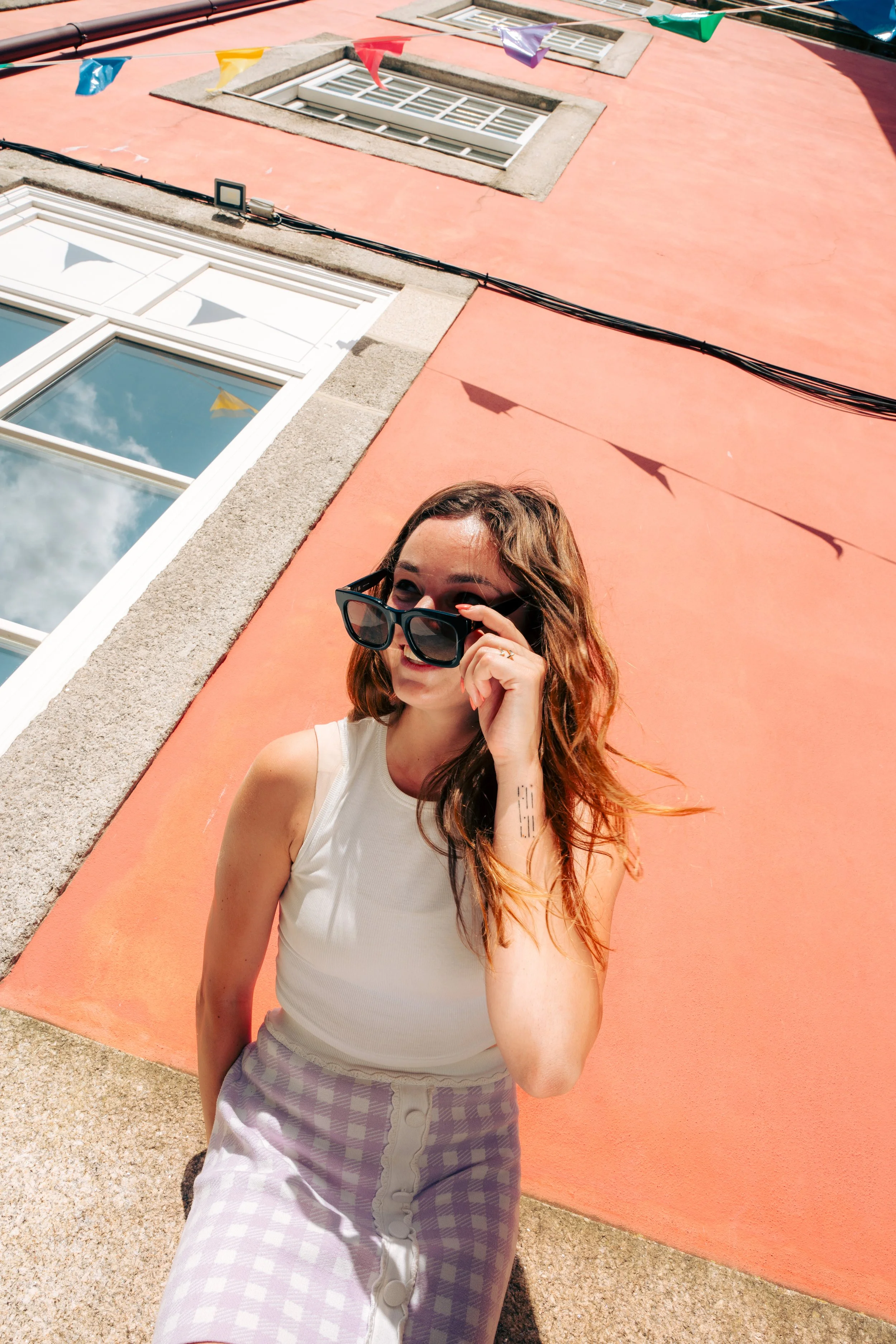 Jeune femme portant des lunettes de soleil, chemise blanche et pantalon à carreaux, se tenant devant un mur de couleur saumon, avec une fenêtre et des drapeaux colorés suspendus à une corde au dessus de sa tête, sous un ciel en partie nuageux.