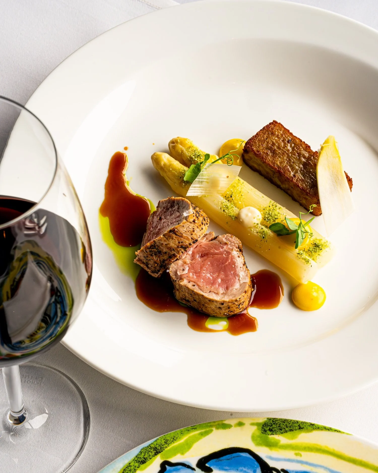 Assiette avec un morceau de viande en croûte, un filet de poisson, des asperges, des sauces et des garnitures végétales, accompagnée d'un verre de vin rouge.