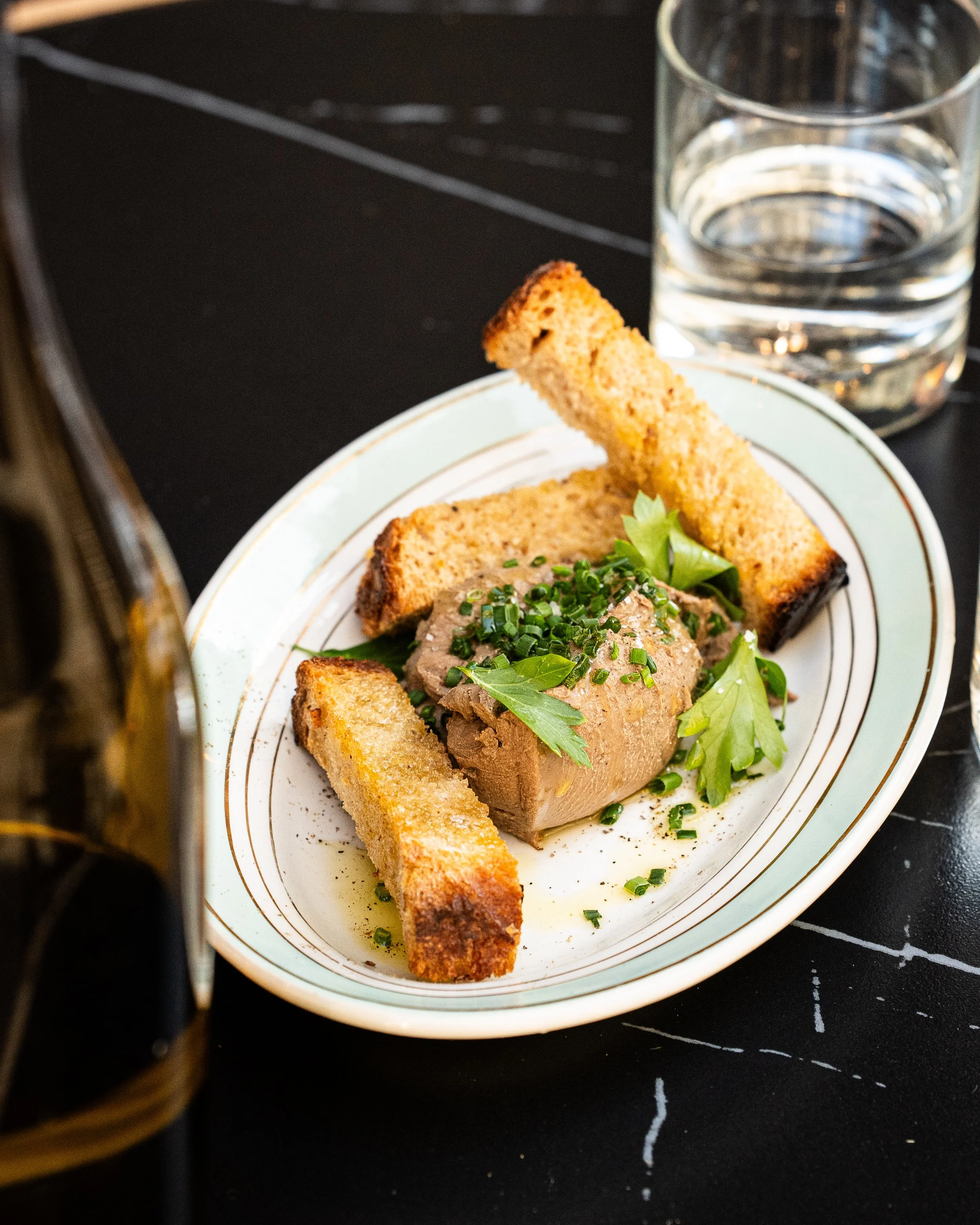 Une assiette blanche avec une tranche de pâté garnie de ciboulette et de feuilles de basilic. À côté, un toast croustillant. Un verre d'eau clair à côté de l'assiette.