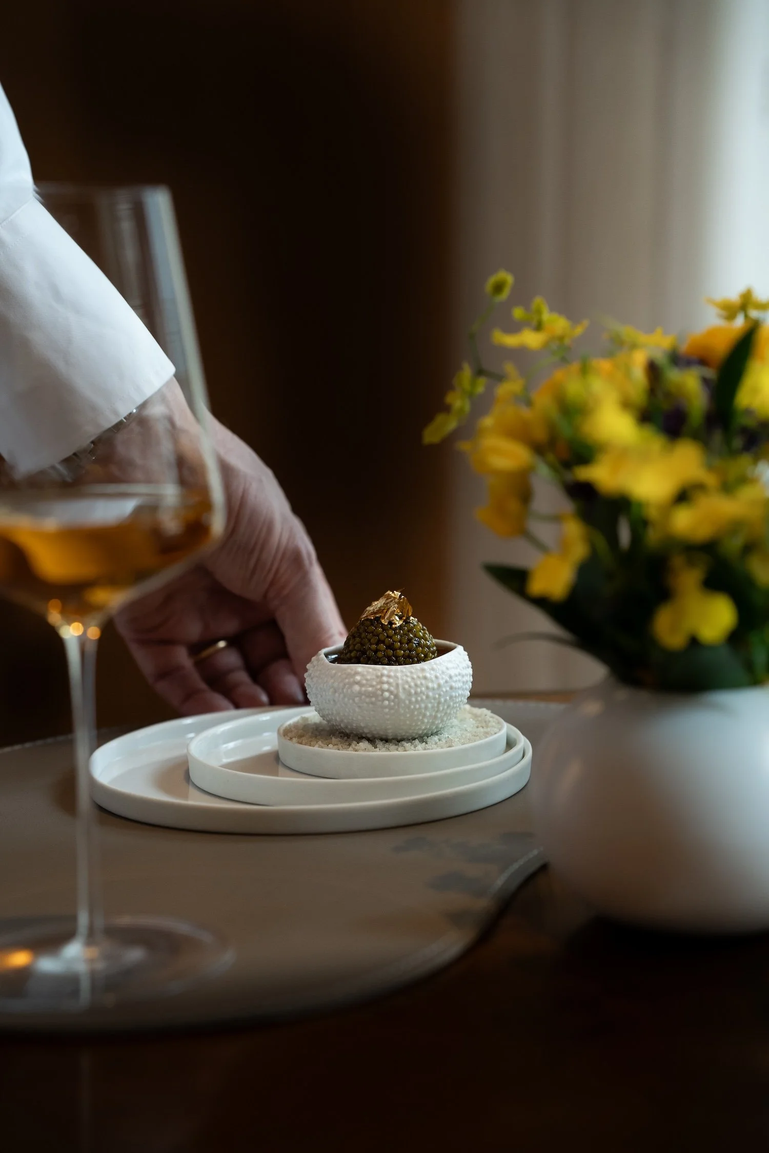 Une main verse un liquide dans une petite coupe contenant une boule faite de petits morceaux, avec un bouquet de fleurs jaunes en arrière-plan.