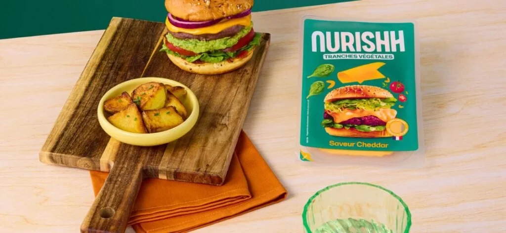 Burger végétarien avec plusieurs tranches de légumes, accompagné de potatoes dans un petit bol, sur une planche en bois, à côté d'une boîte de fromage végétal et d'un verre vide, sur une table en bois claire.