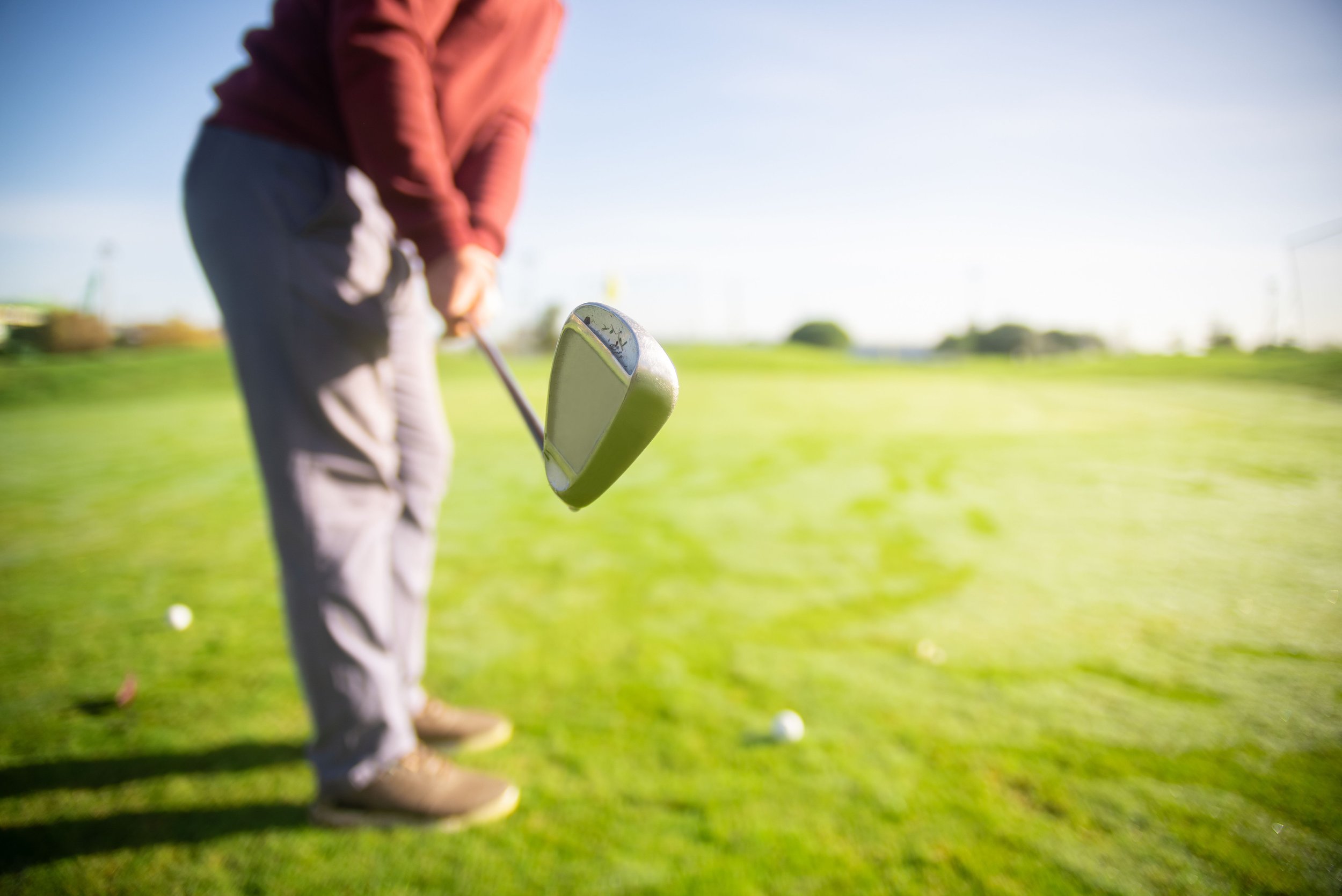 A person preparing to hit a golf ball on a golf course, holding a golf club with a focus on the club head, with a bright sky and green grass in the background.