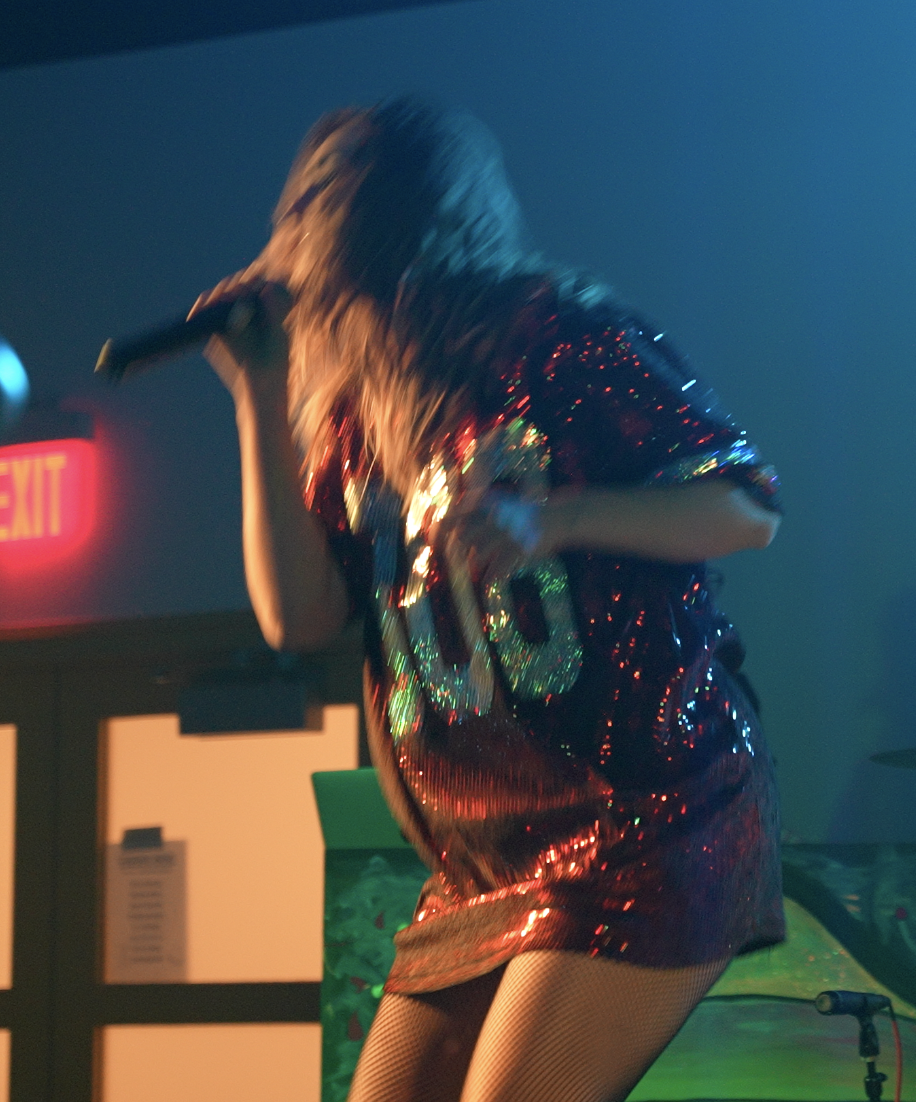 Female singer performing on stage with a microphone, dressed in a shiny, sequin-covered dress, with colorful stage lighting in the background.