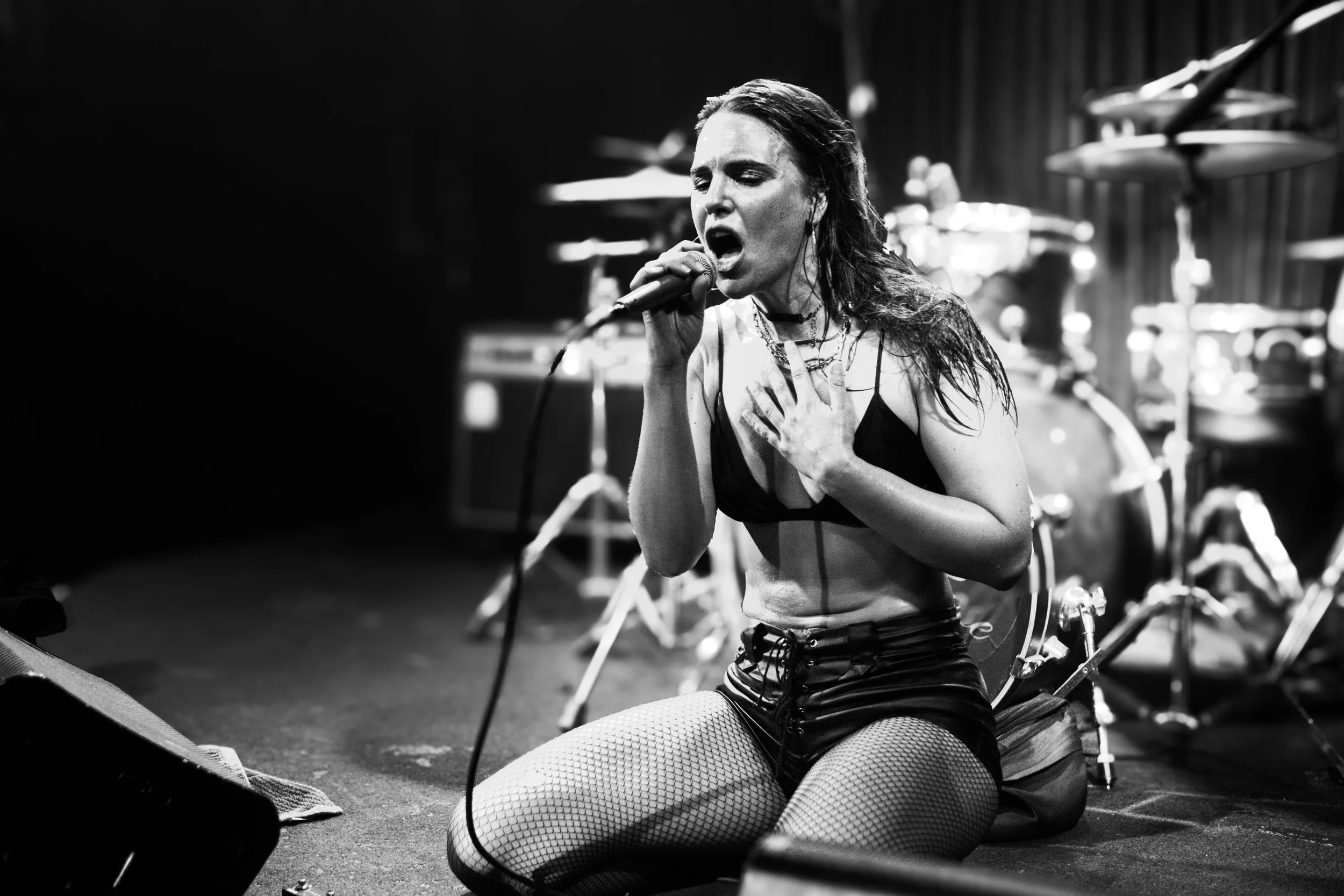 A black and white photo of a woman singing with her eyes closed, one hand on her chest, and the other holding a microphone. She appears emotional and is wearing fishnet stockings and a sleeveless top, with a drum set in the background.