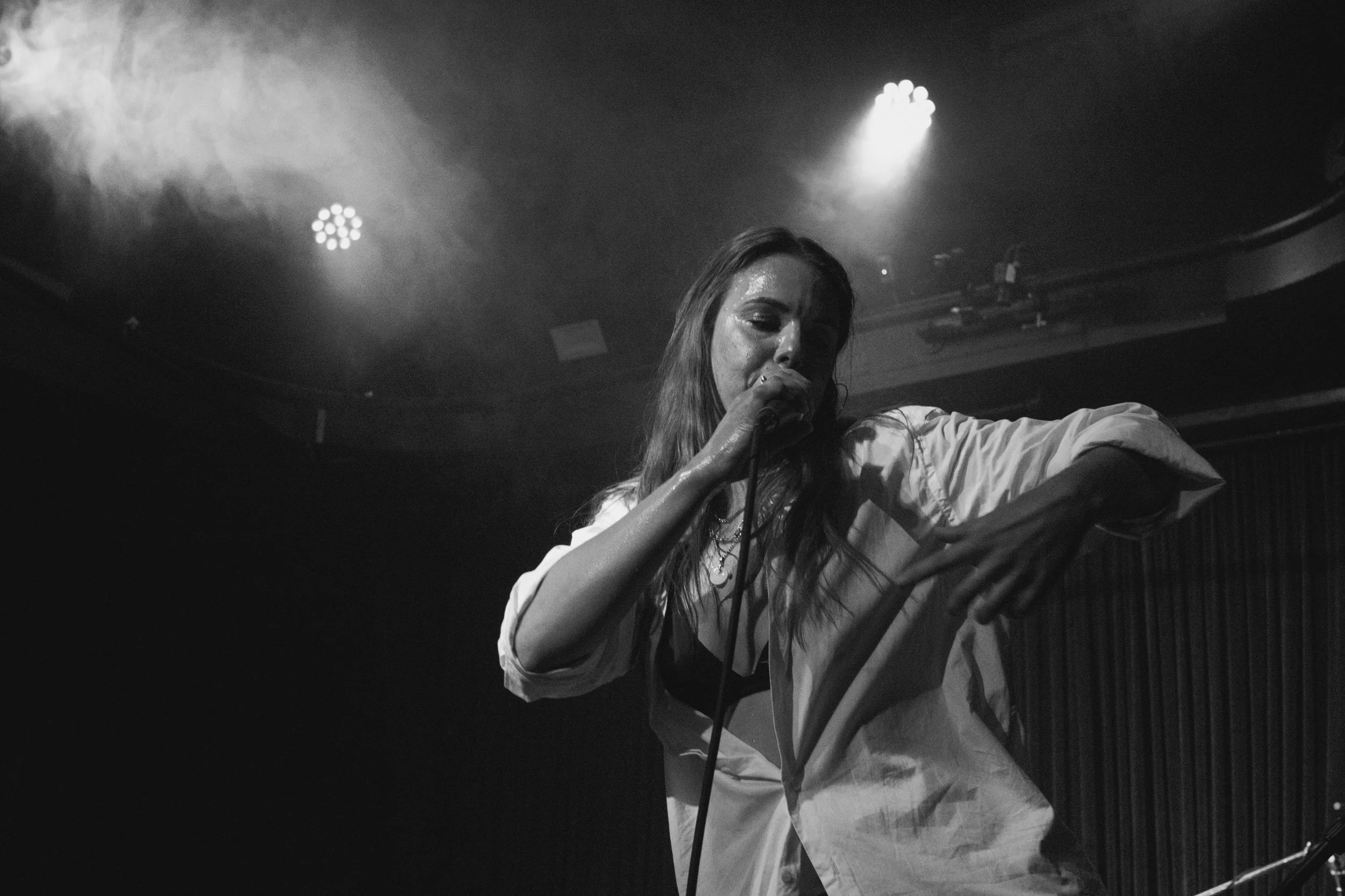 A woman performing on stage with a microphone, wearing a light-colored shirt, in a dark auditorium with stage lights overhead.