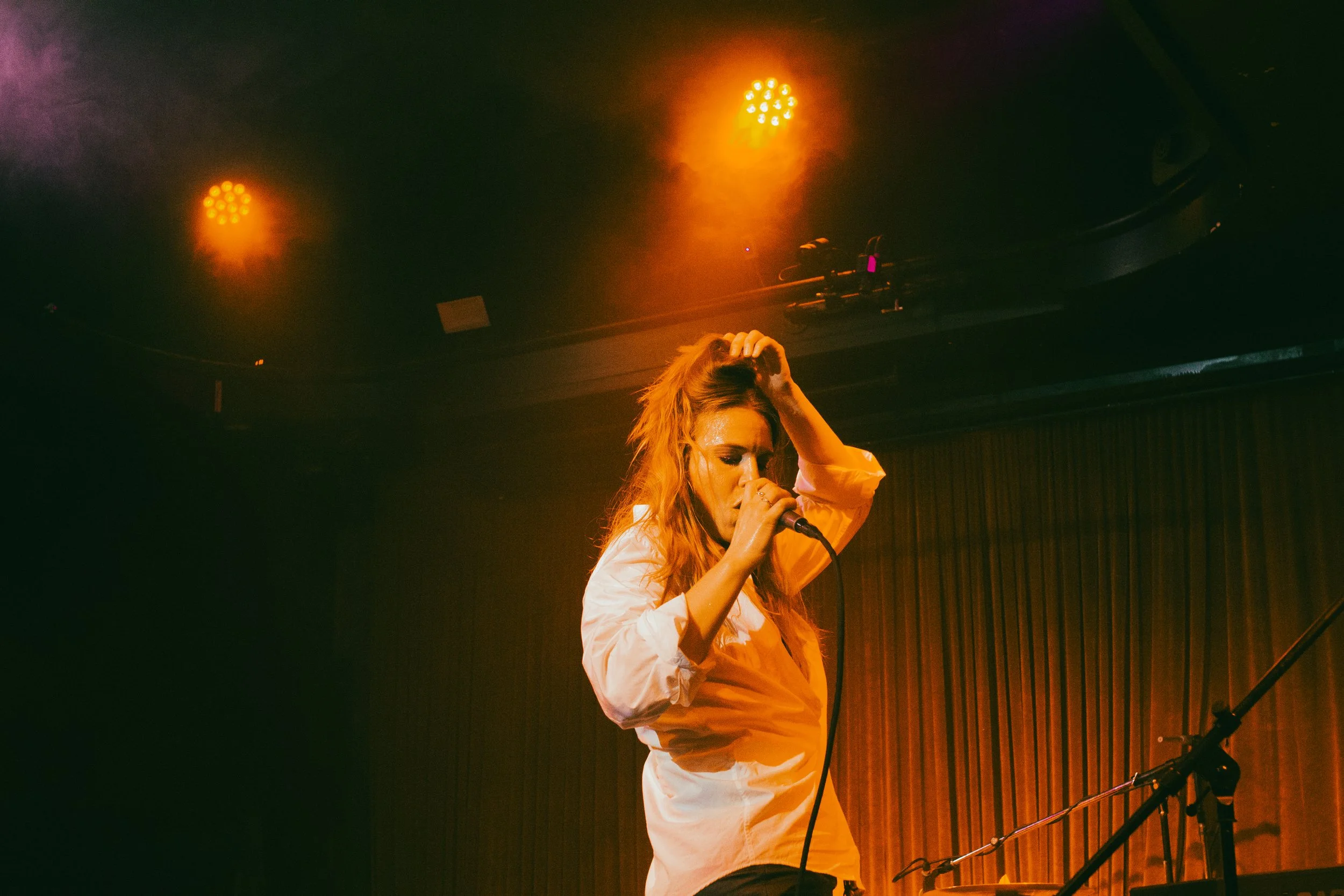 A woman with long hair singing into a microphone on stage with orange and purple stage lights overhead.