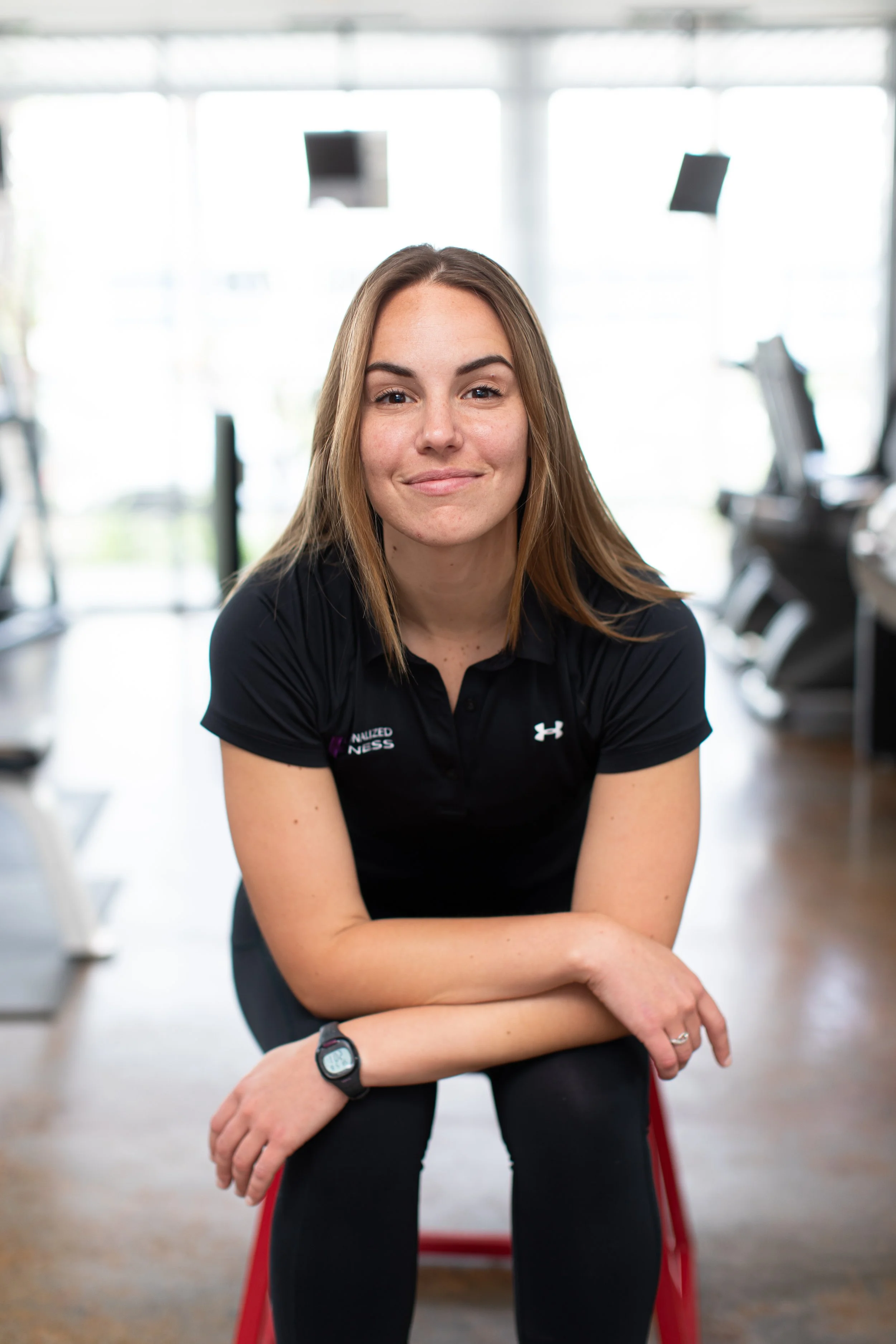 A young woman with brown hair and a black Under Armour shirt sitting on a red workout bench inside a gym, smiling at the camera with a fitness tracker on her left wrist.