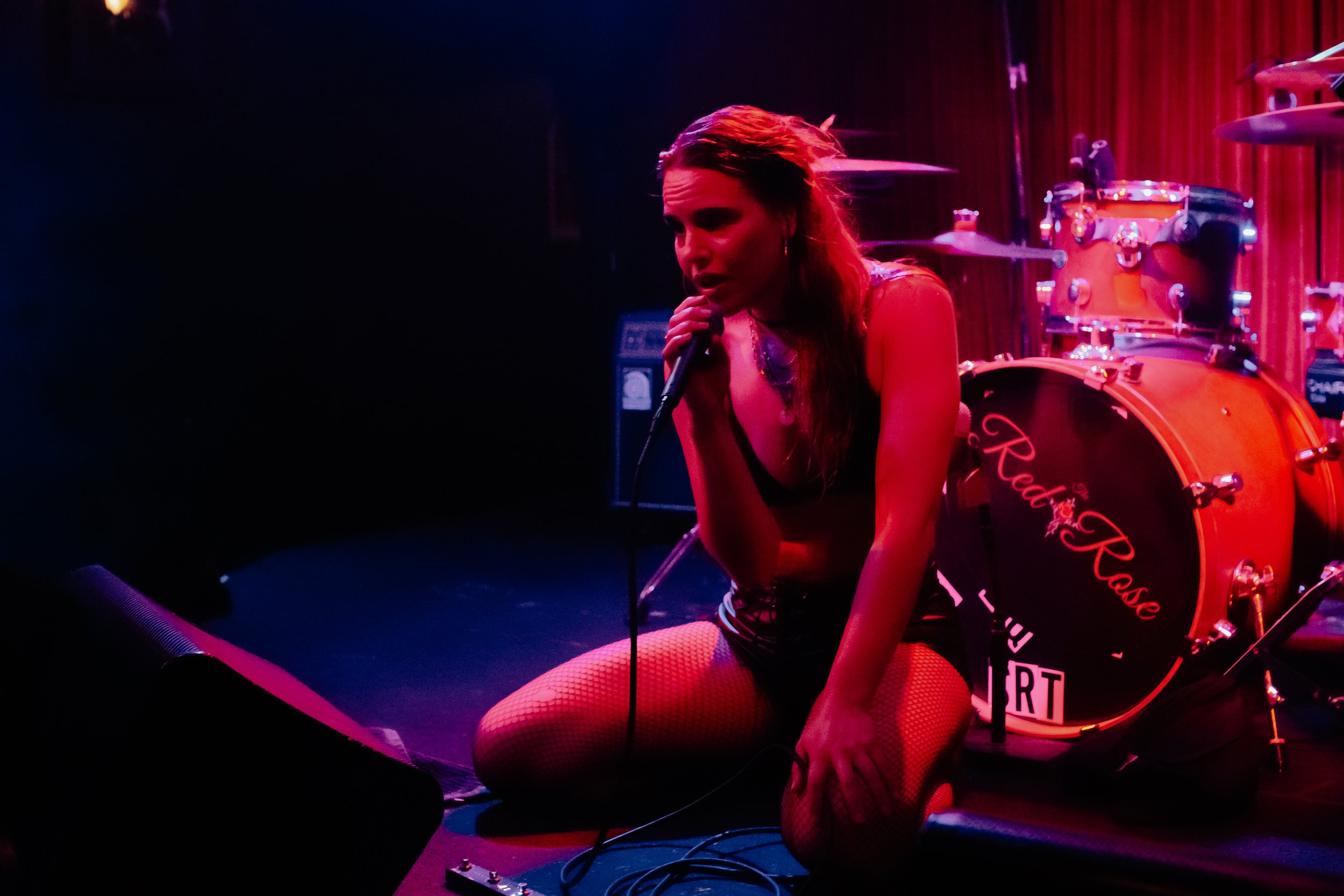 A woman singing into a microphone on stage, with stage lights casting a pink and purple hue, and a drum set in the background.