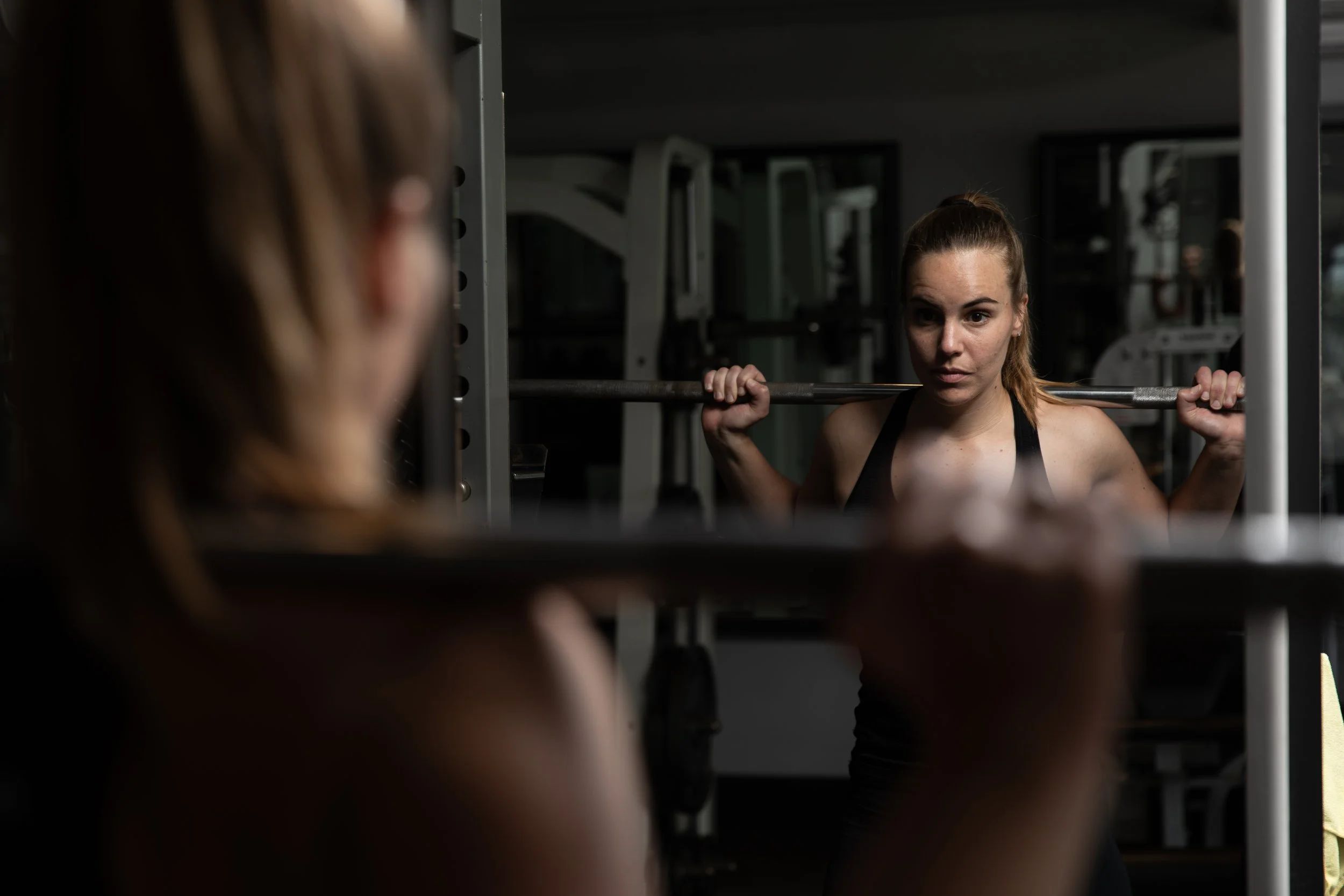 A woman is lifting weights in a gym while looking at herself in a mirror.