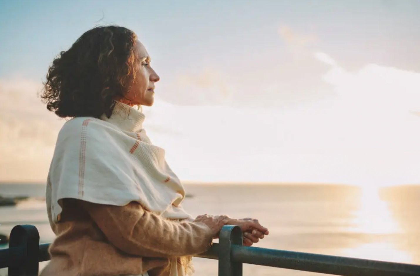 A woman with curly hair looking at the sunset over the ocean while leaning on a railing.