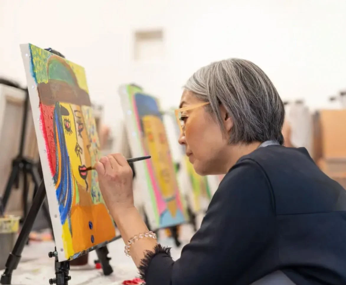 A woman with gray hair and glasses painting a colorful abstract portrait on a canvas in an art studio.