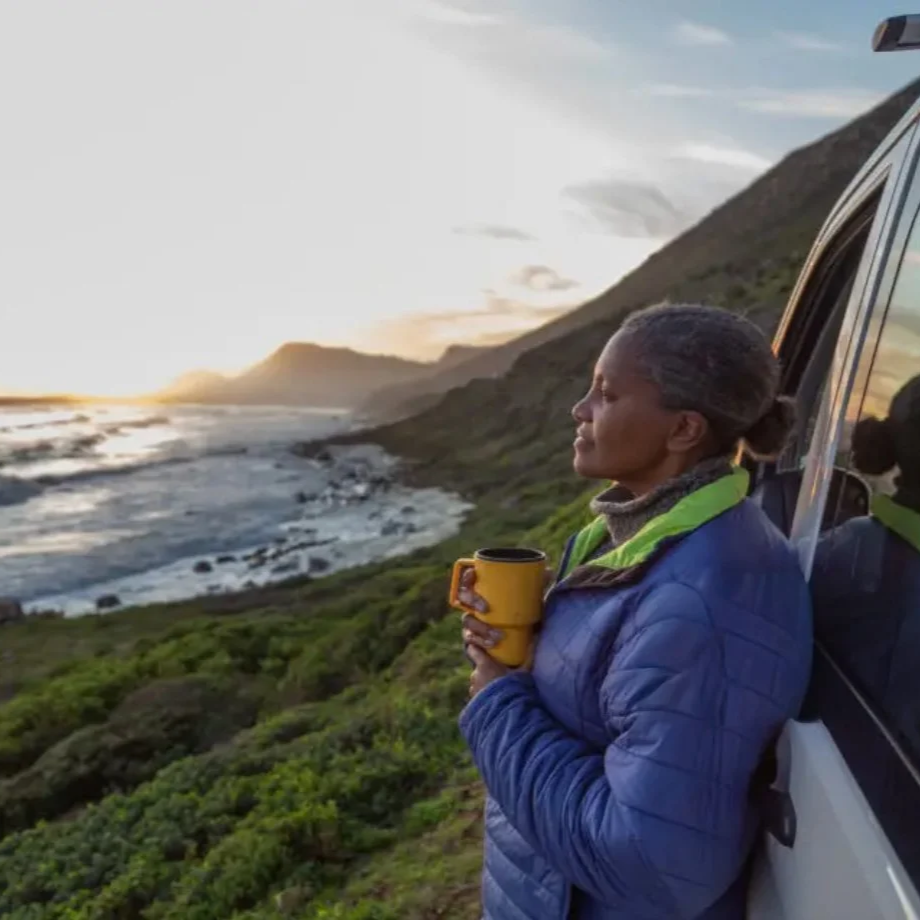 An elderly woman with gray hair standing by an open car door, holding a yellow mug, gazing at a coastal landscape with hills, rocky shoreline, and waves during sunset.