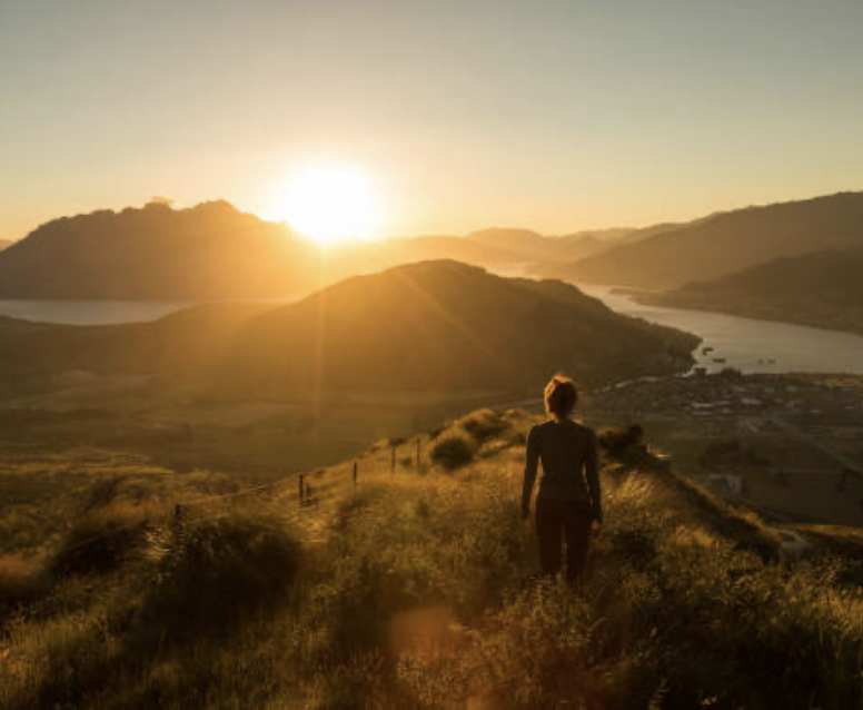 Person walking along a hilltop during sunset with mountains and a river in the background.