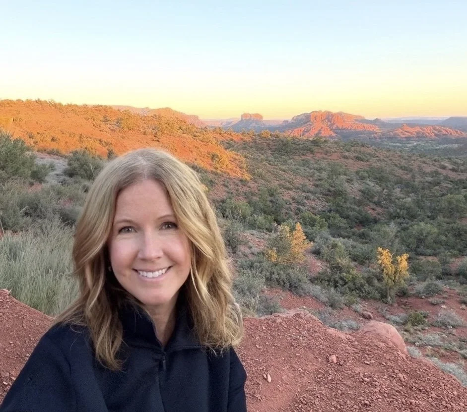 A woman with blonde hair smiling at the camera in a desert landscape at sunset, with red rocks, sparse green vegetation, and distant mesas.