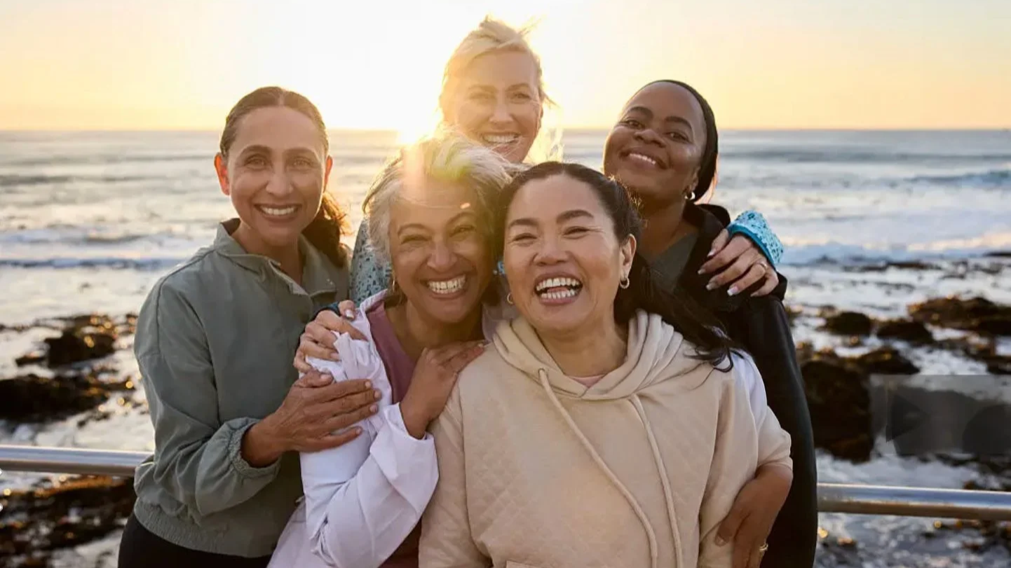 Five women smiling and hugging each other at the beach during sunset.