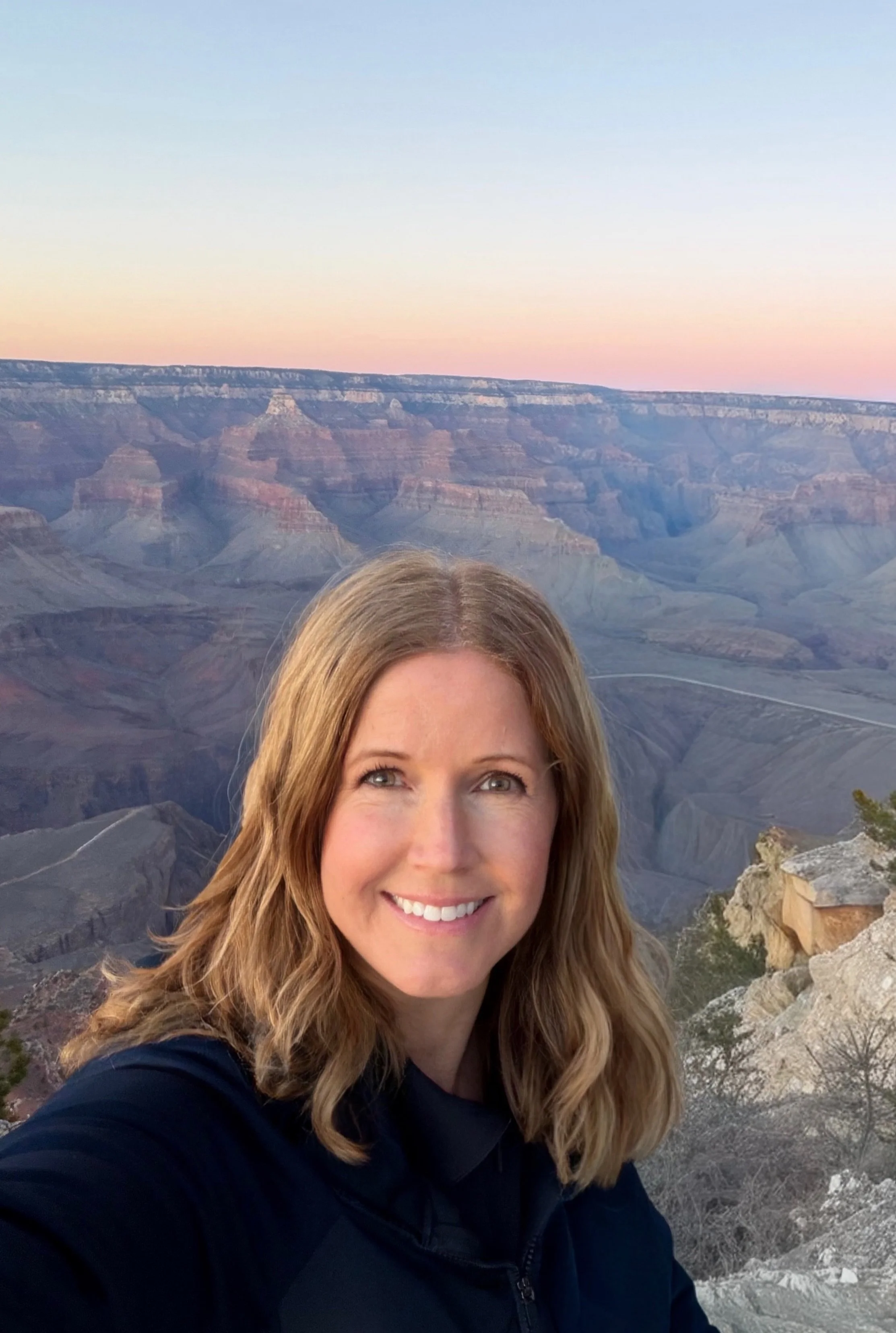 A woman with long wavy blonde hair smiling, standing near the Grand Canyon during sunset with colorful sky and canyon landscape in the background.