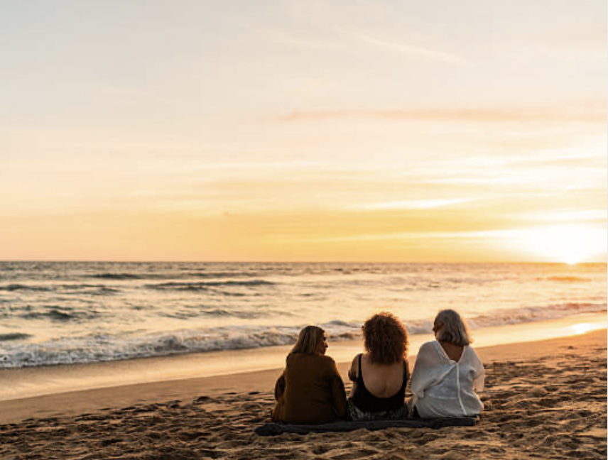 Three women sitting on the beach at sunset, facing the ocean.
