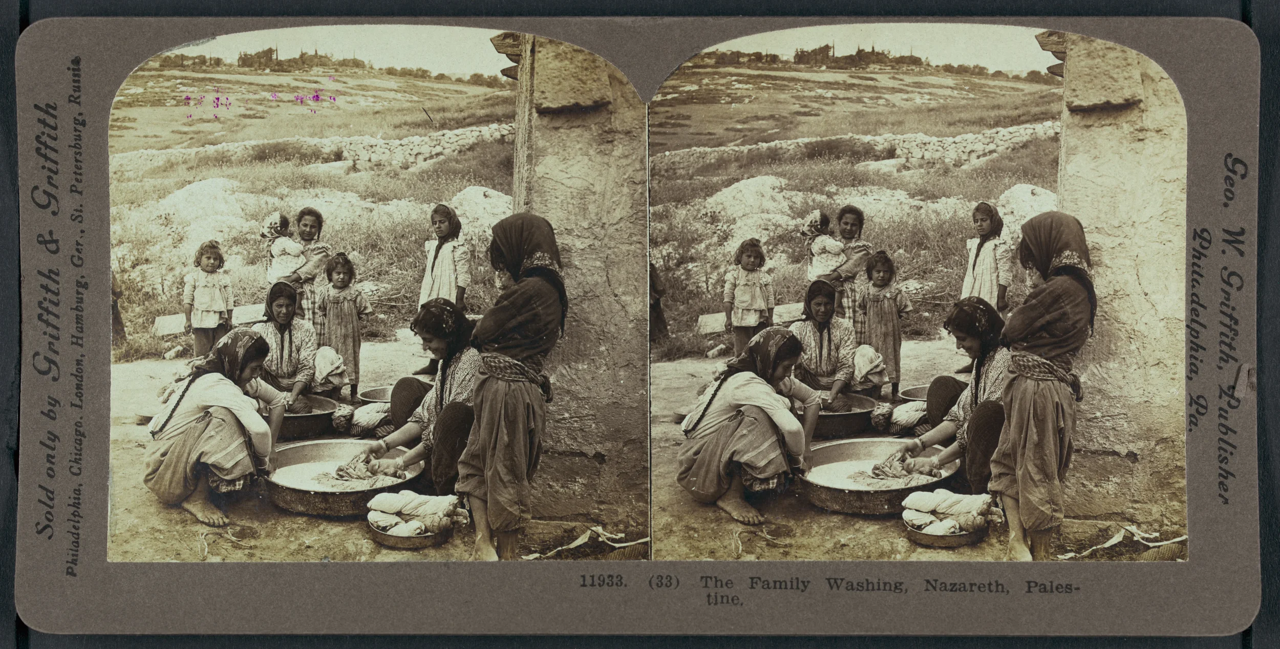 Historic black-and-white photograph of a family washing clothes in Nazareth, Palestine, with women and children gathered around large basins, outdoors near a stone wall and open landscape.