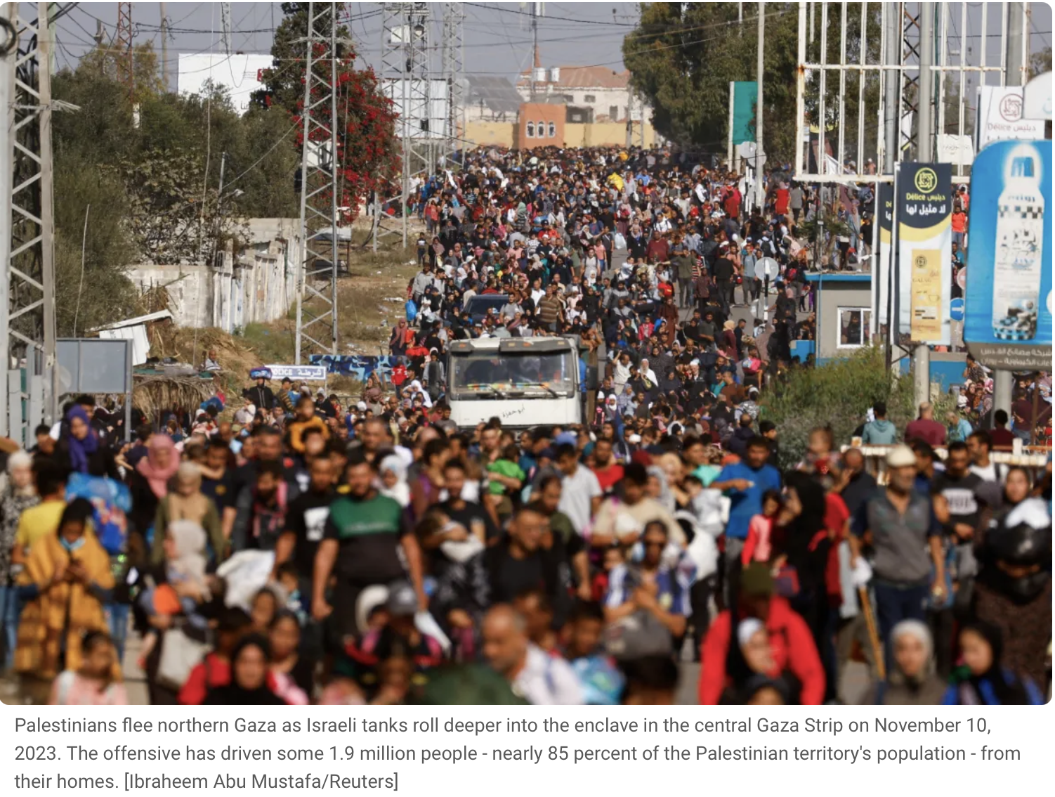 Large crowd of Palestinians fleeing northern Gaza on a busy street, with power lines and vehicles, as Israeli tanks roll into the area in the background.