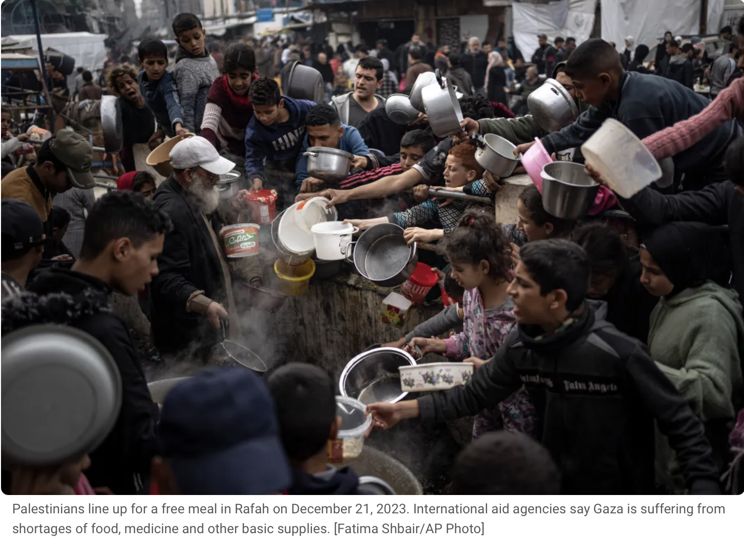 Palestinians line up for a free meal in Rafah, with many people reaching out for food from a large cooking pot, as others stand behind waiting.