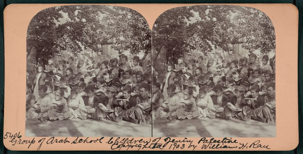 Black and white photograph of a large group of Arab school children sitting outdoors under trees, reading books. The photo was taken in 1903 by William H. Rau.
