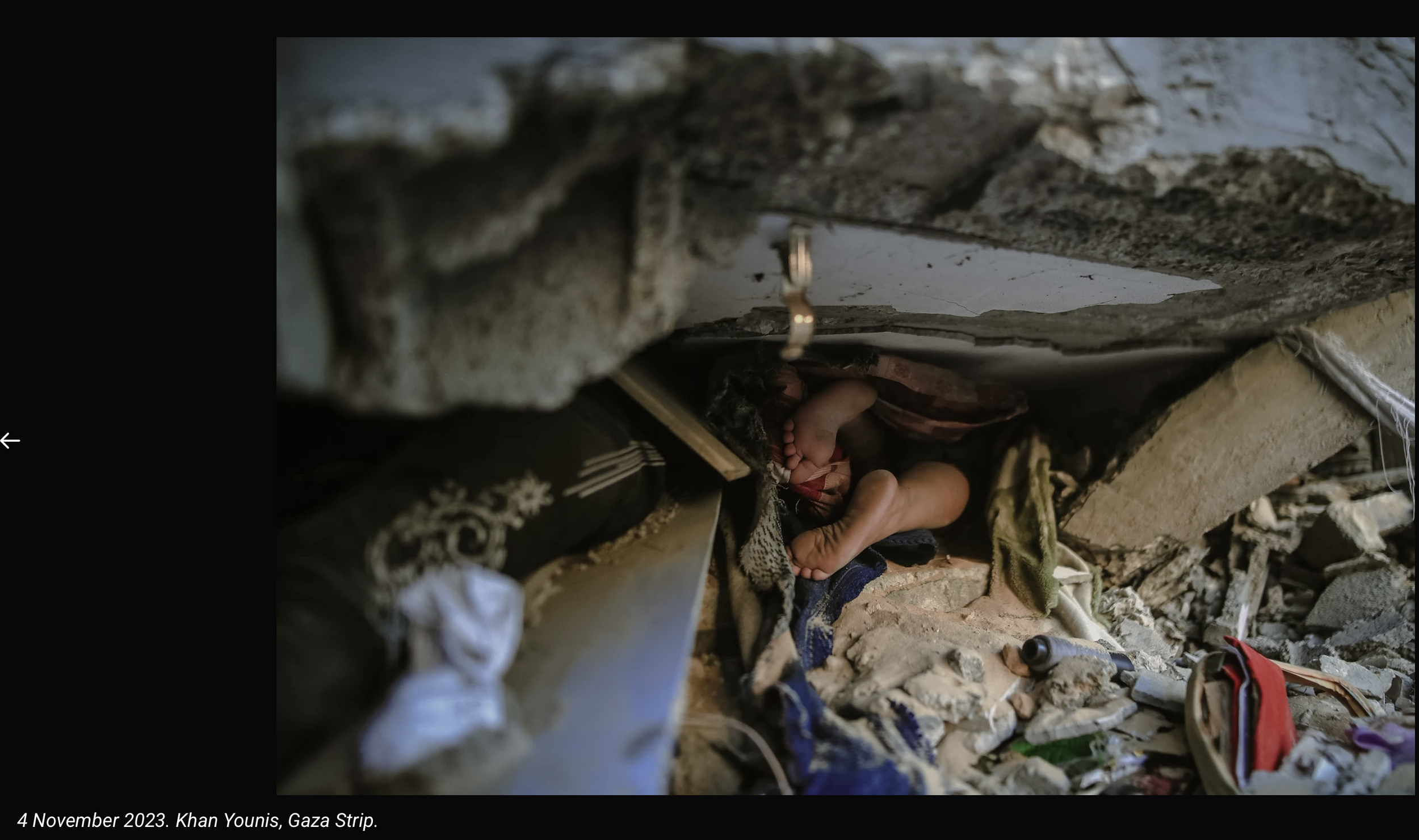 A narrow, rubble-filled alleyway in Gaza Strip with debris, trash, and broken objects, including a child lying on the ground, seen from a low angle.
