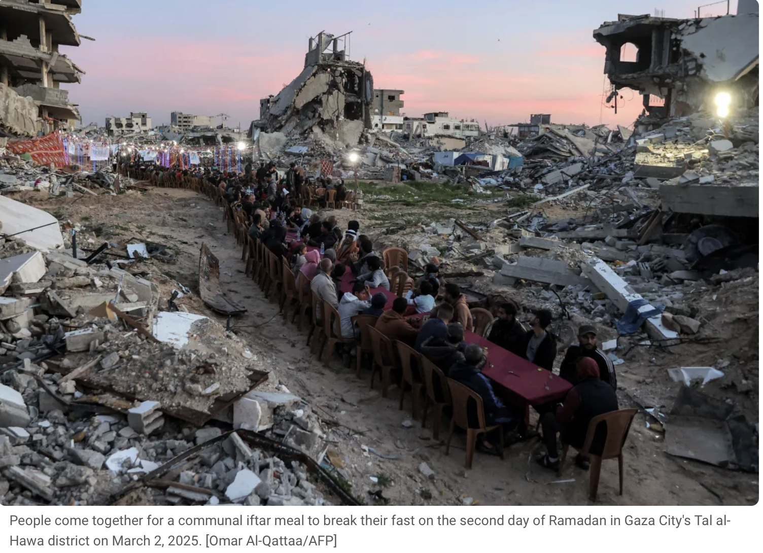 People are seated at long tables outdoors, celebrating an iftar meal amidst destruction and rubble from buildings that have been heavily damaged or destroyed in Gaza City.