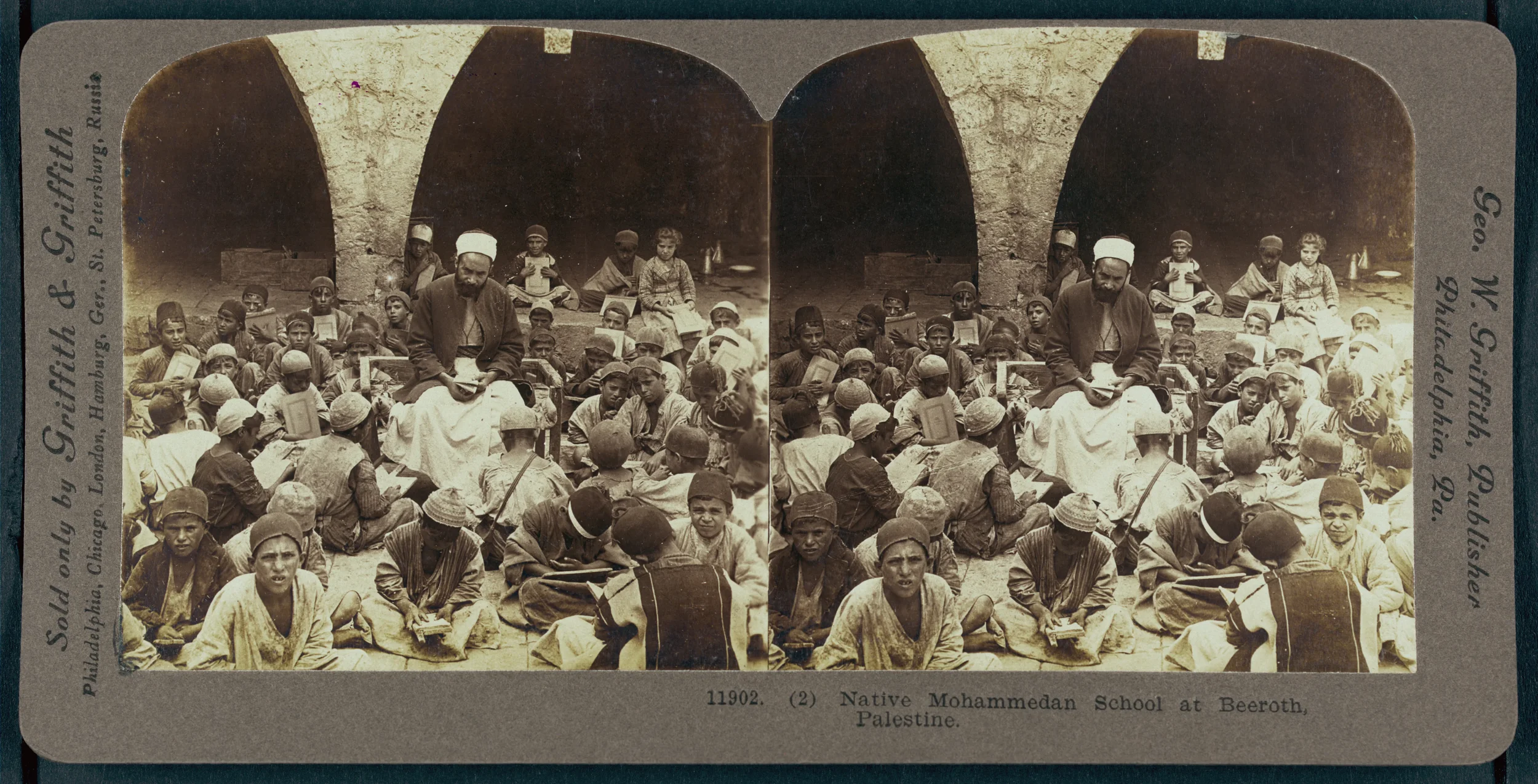 Historical black and white photograph of a large group of children and a few adults seated outdoors in front of a stone structure with arches. The scene depicts a classroom setting at the Native Mozambican School at Beeroth, Palestine, with children 