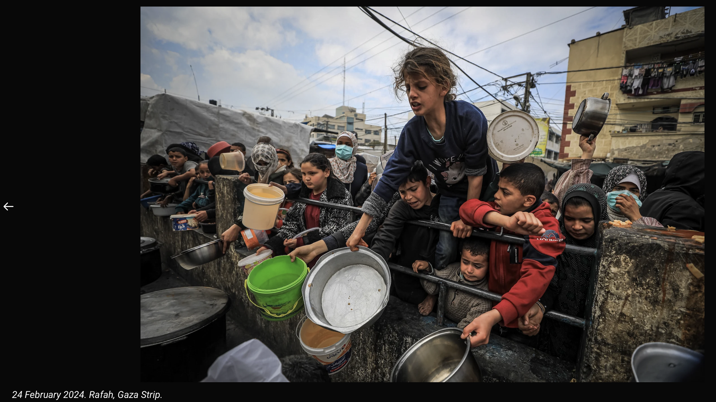 Children lining up to receive food outdoors on Gaza Strip street in Rafah, showing many with empty pots and plates, amid damaged buildings and overhead power lines.