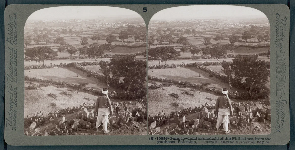 A valley with scattered trees and cultivated fields, seen from a hillside. In the foreground, a man in traditional Palestinian attire, including a white head covering, stands among a group of seated villagers. This is a historical postcard image of G